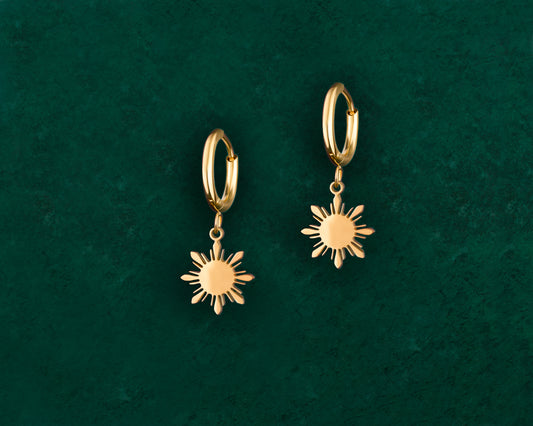 A pair of Aking Sinta gold Araw charm dainty huggie earrings on a lush green background, featuring small gold hoops with suspended 8-ray sunburst charms.