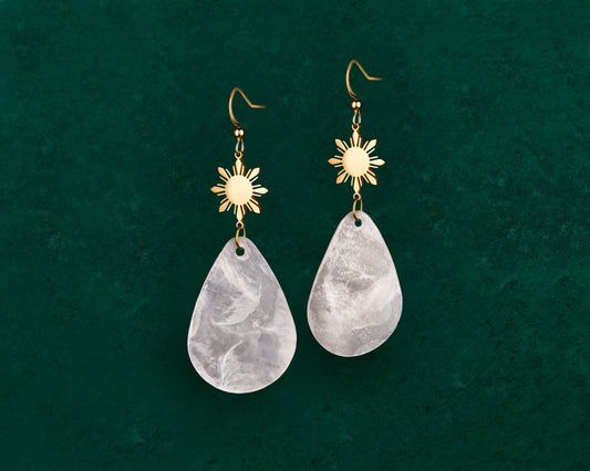 A pair of Aking Sinta gold hook earrings featuring a polished 8-ray Araw sun charm at the top, with a dangling iridescent teardrop-shaped Capiz shell suspended below, displayed on a vibrant green background.