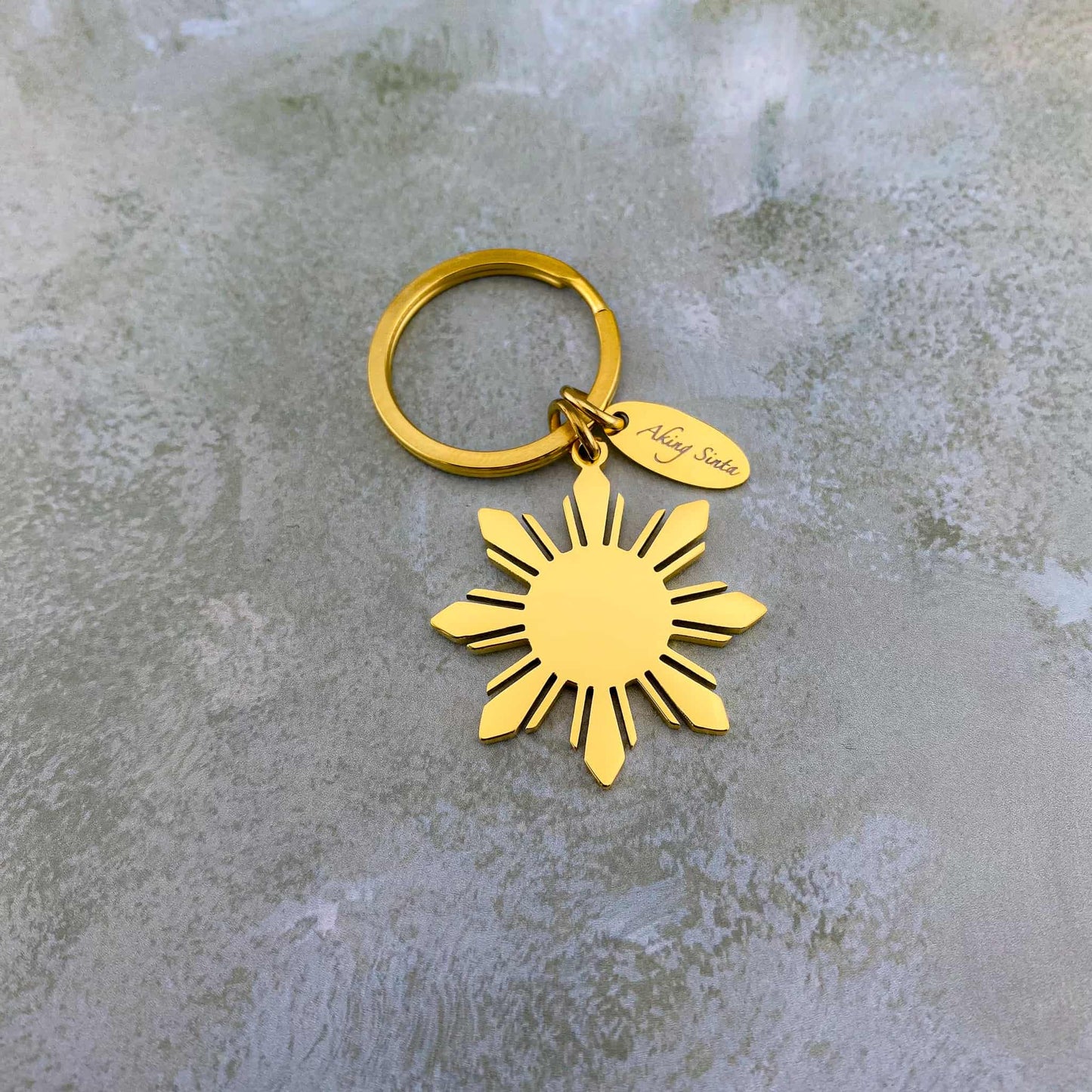 The gold Aking Sinta Araw key ring featuring a large 8-ray sunburst charm, displayed on a sleek gray marble background to highlight the radiant polished finish of the gold plating