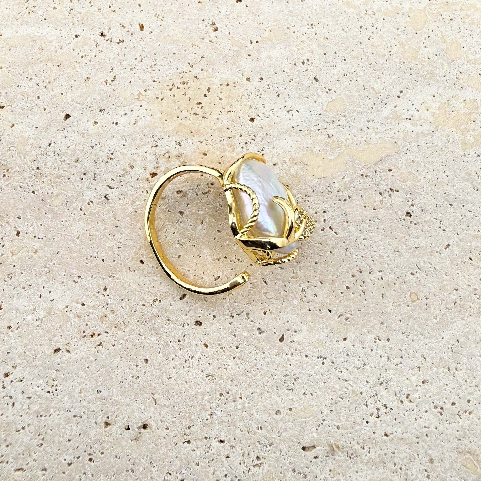 A close-up view of an adjustable Aking Sinta gold ring, highlighting the open-back band design. The ring features a lustrous Keshi pearl encased in decorative gold wire with a small, sparkling gold leaf detail, resting on a light, textured natural stone surface