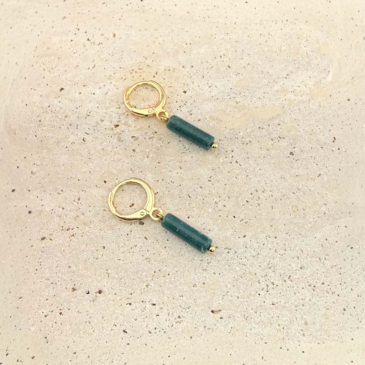 A pair of Aking Sinta gold huggie hoop earrings featuring dark green African jade column pendants. These natural gemstone drop earrings are displayed on a light, textured stone surface.