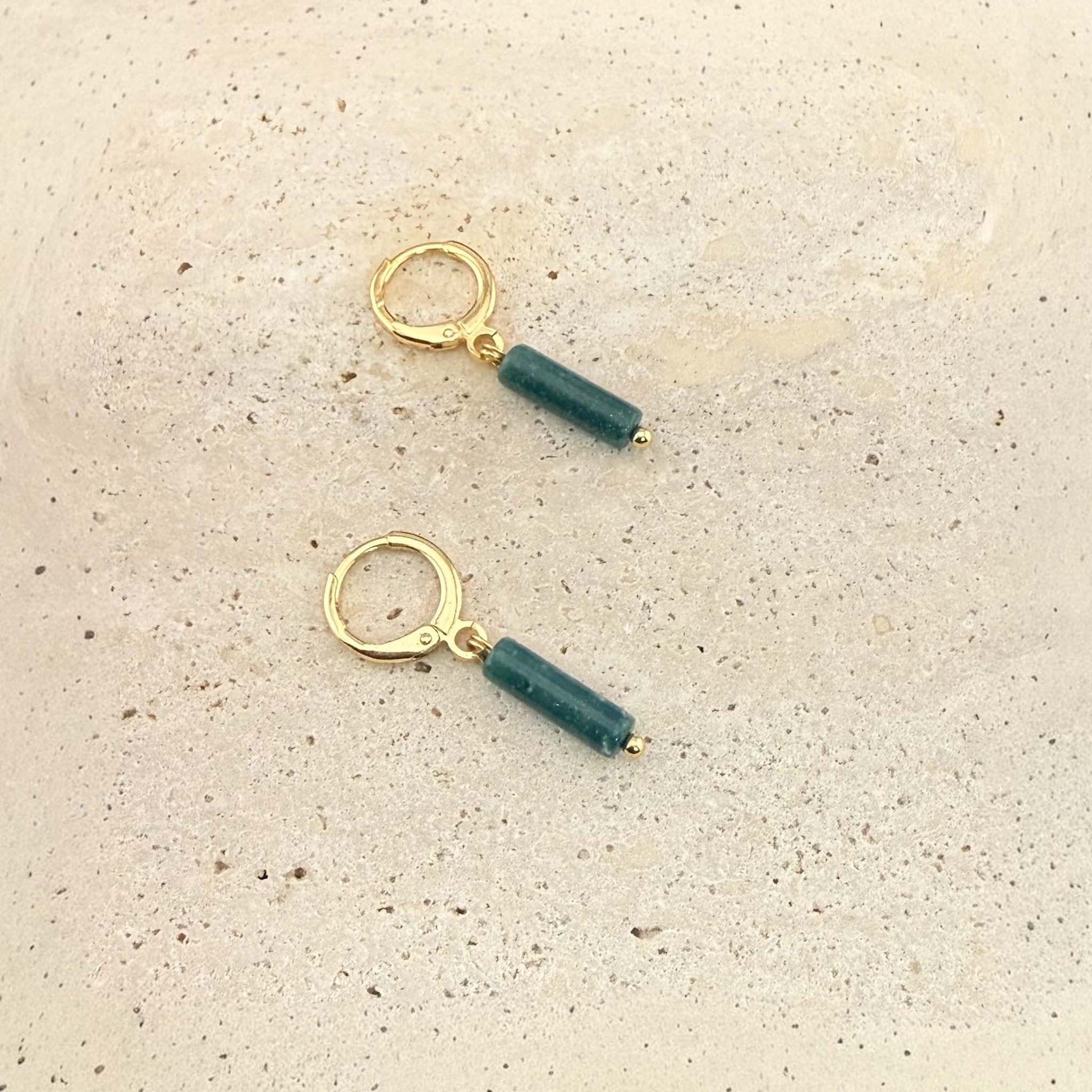 A pair of Aking Sinta gold huggie hoop earrings featuring dark green African jade column pendants. These natural gemstone drop earrings are displayed on a light, textured stone surface.