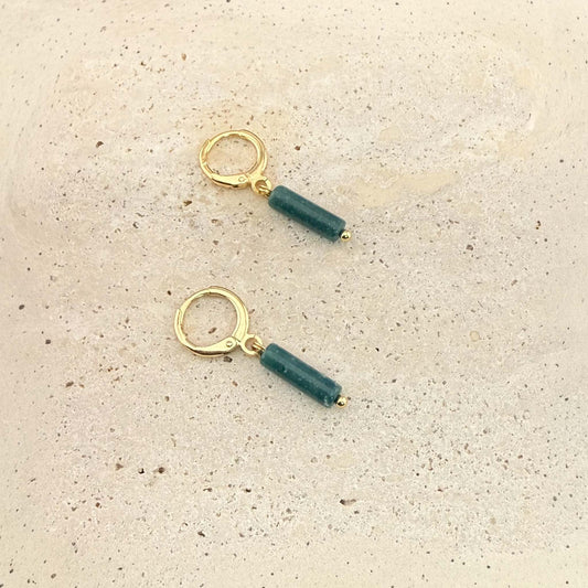 A pair of Aking Sinta gold huggie hoop earrings featuring dark green African jade column pendants. These natural gemstone drop earrings are displayed on a light, textured stone surface.