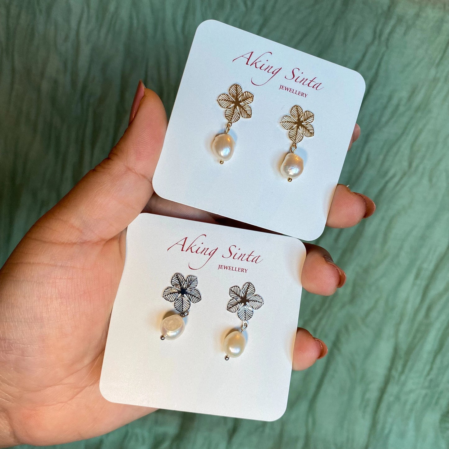 A hand holding two pairs of Aking Sinta statement earrings presented on white branded display cards. The top pair features gold filigree Sampaguita flower studs with baroque pearl drops, while the bottom pair features silver-tone Sampaguita flower studs with matching pearl drops. The hand is set against a soft, textured green background