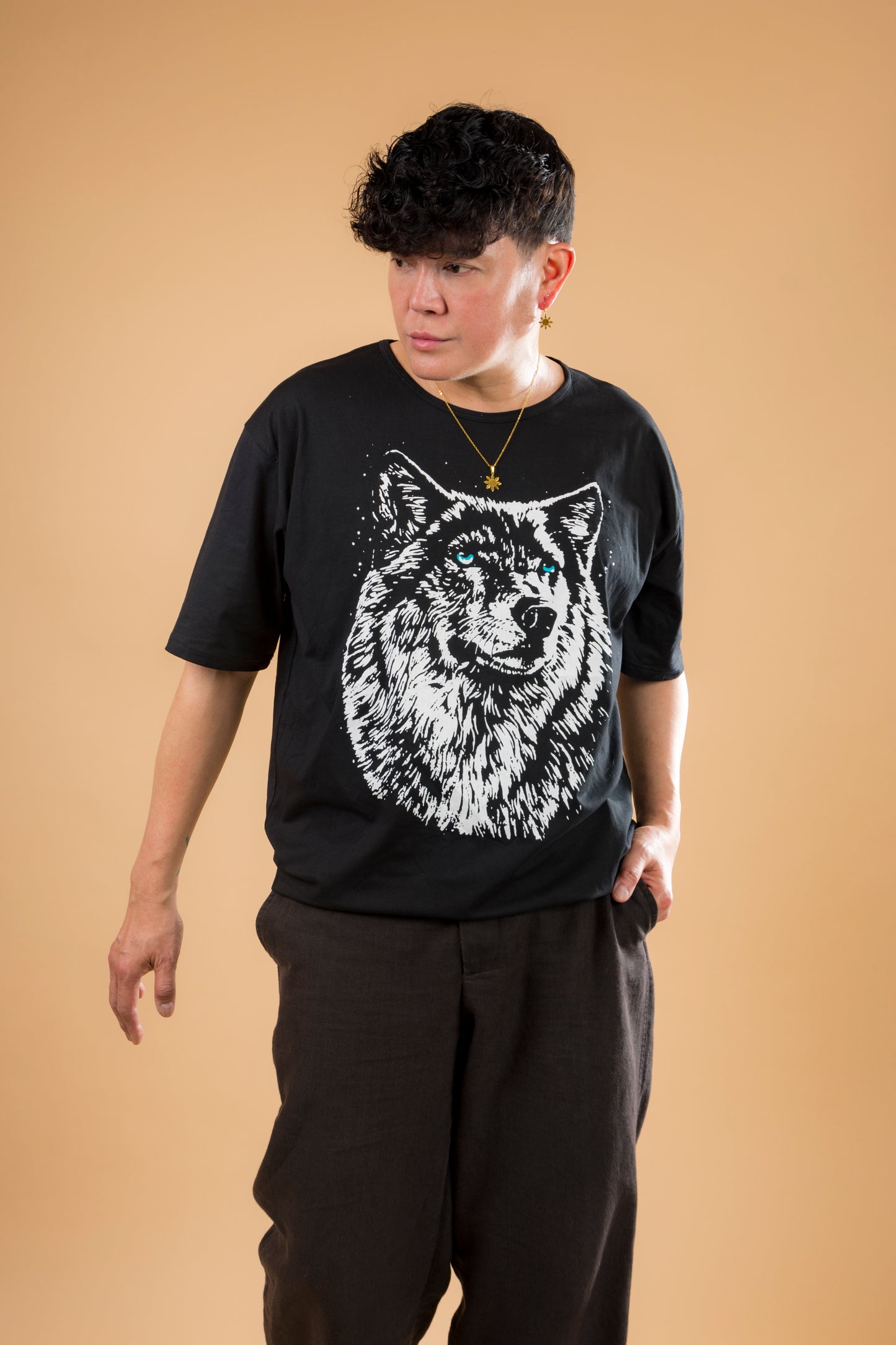 A full-body lifestyle portrait of a man wearing the Aking Sinta gold Araw necklace and matching sun stud earring. Styled with a graphic wolf t-shirt and dark trousers against a neutral tan studio backdrop, the image highlights the unisex and contemporary appeal of Filipino-inspired heritage jewelry.