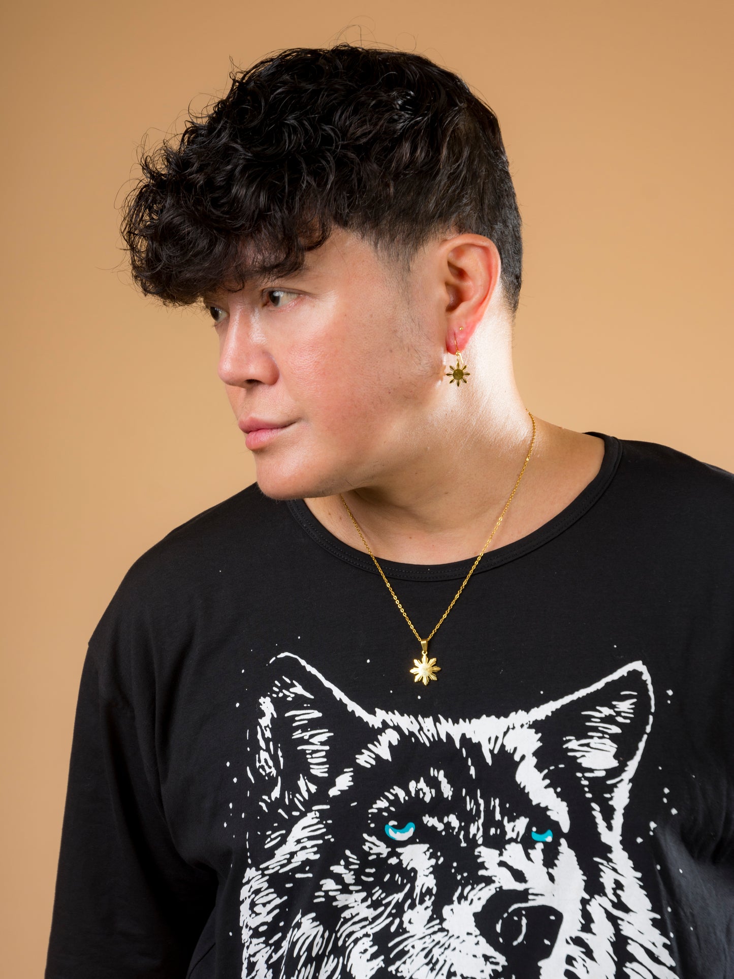A studio lifestyle portrait of a man wearing the Aking Sinta gold Araw necklace and matching Araw drop earring. The eight-rayed Philippine sun jewelry is styled with a graphic black t-shirt against a neutral tan background, showcasing the collection's modern, unisex, and culturally significant design.