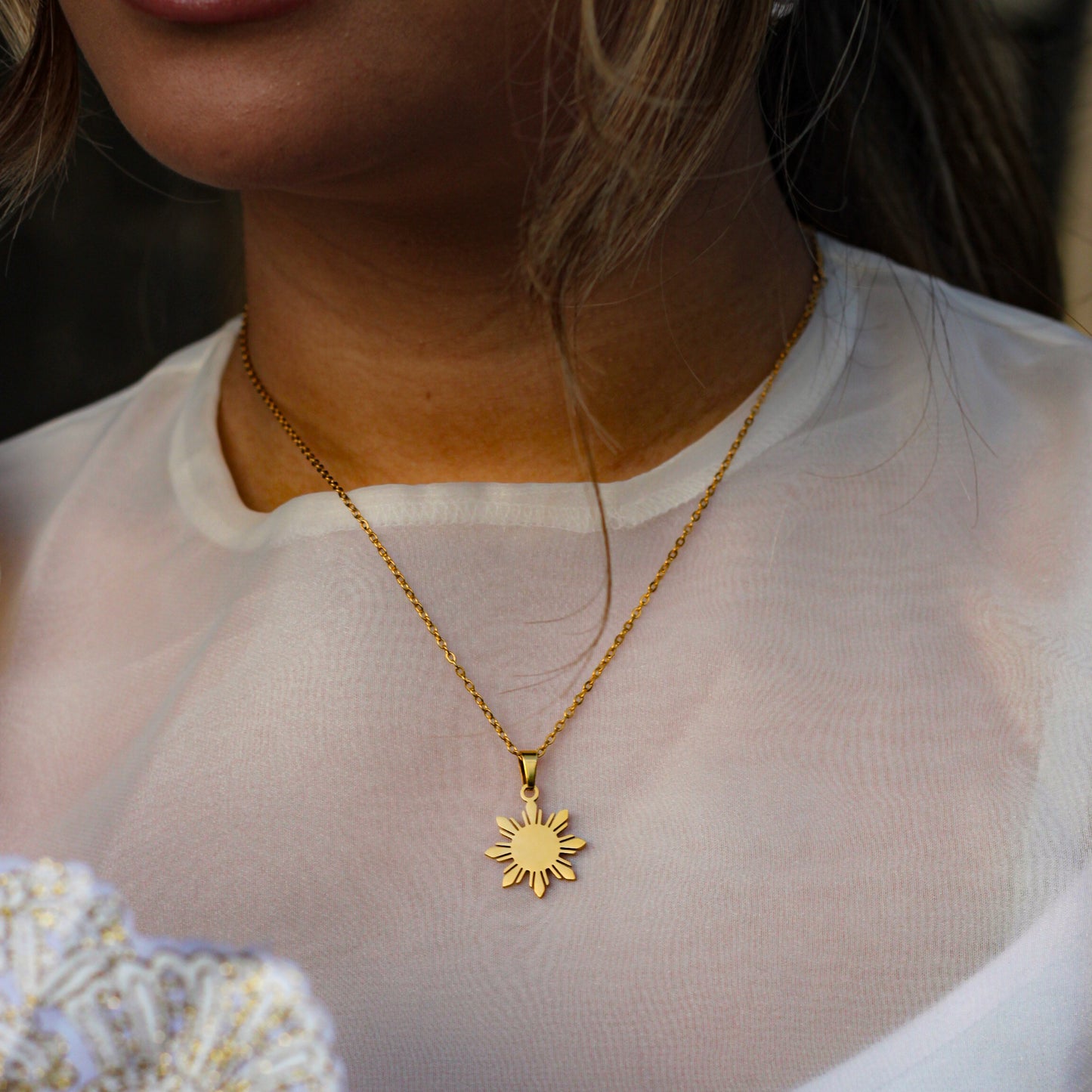 A lifestyle close-up of a person wearing the Aking Sinta gold Araw necklace. The minimalist eight-rayed Philippine sun pendant is elegantly styled over a sheer white blouse, highlighting the warm metallic glow and cultural significance of the jewelry.