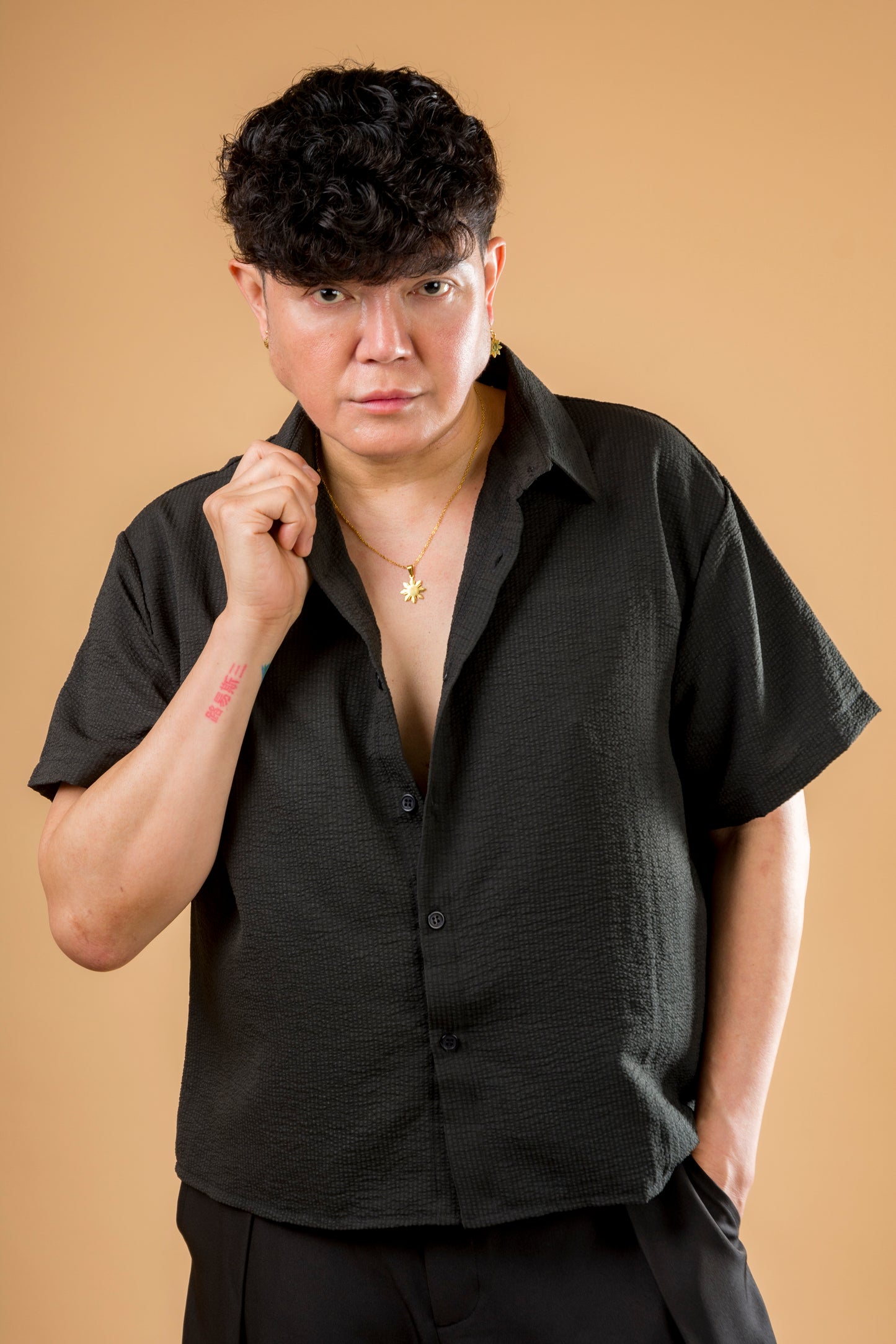A studio lifestyle portrait of a man wearing the Aking Sinta gold Araw necklace. The eight-rayed Philippine sun pendant is styled with a textured black short-sleeve button-down shirt against a warm tan background, demonstrating a sophisticated and modern way to wear Filipino-inspired heritage jewelry.