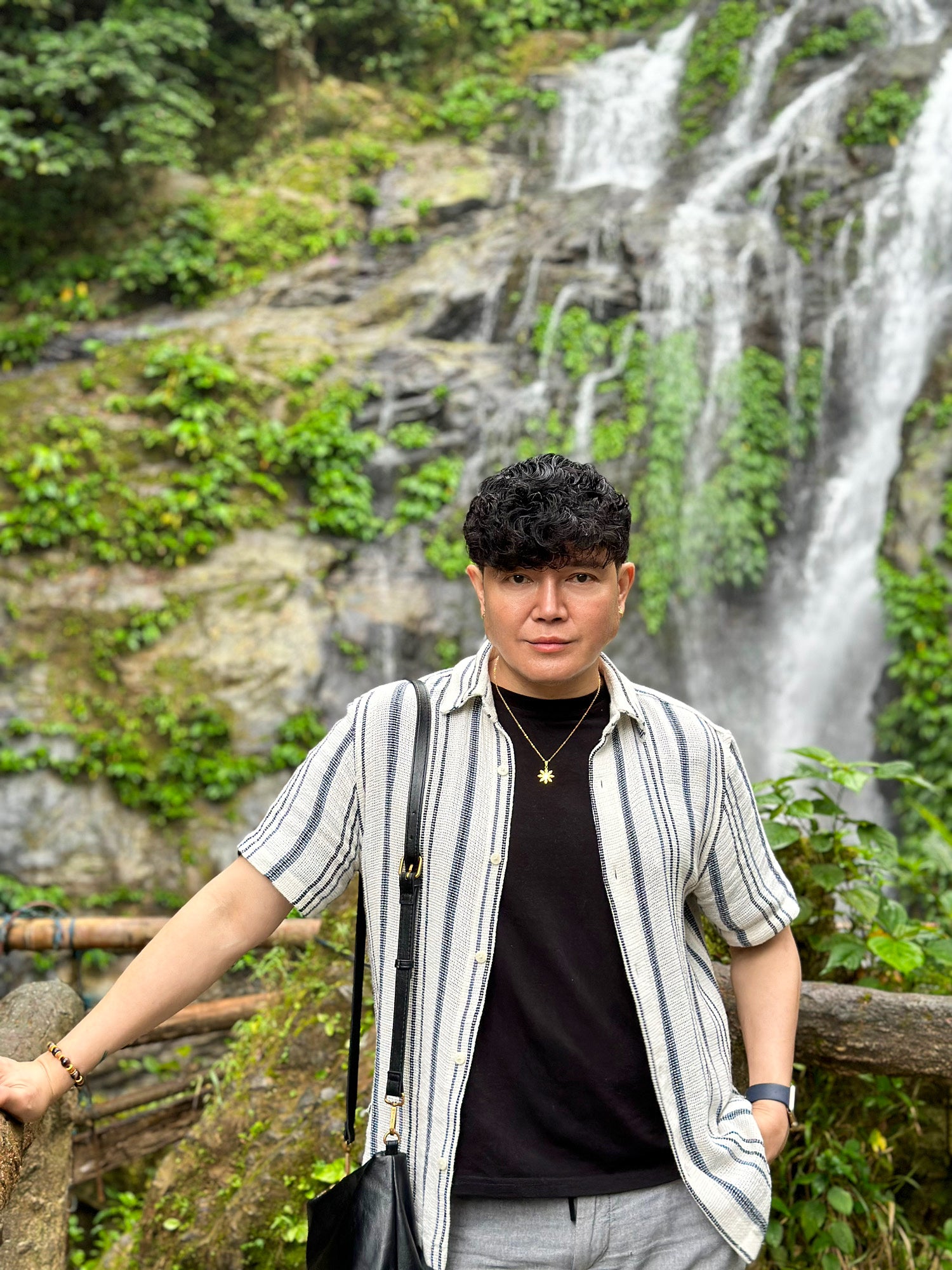 A lifestyle portrait of a man wearing the Aking Sinta gold Araw necklace in an outdoor tropical setting. The minimalist Philippine sun pendant is styled with a casual black shirt and open-striped button-down, set against a lush waterfall background to showcase the piece's versatility and unisex appeal.