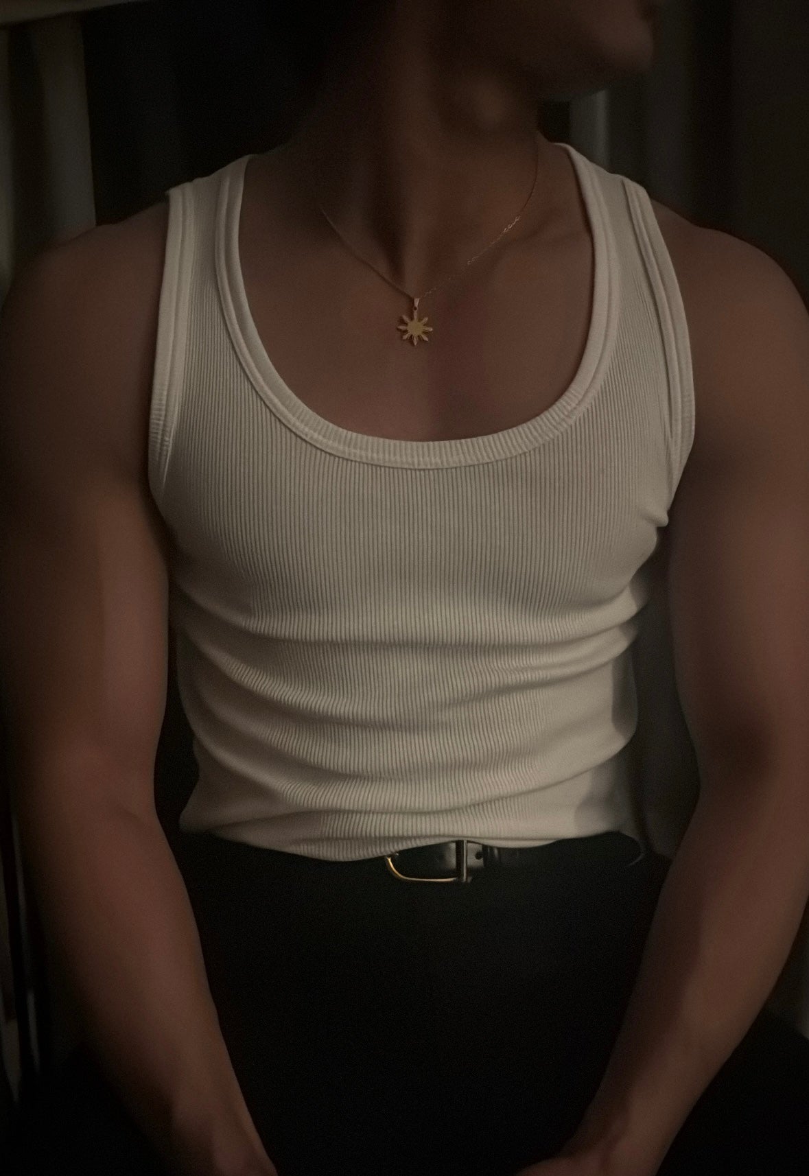 A low-light lifestyle shot of a man wearing the Aking Sinta gold Araw necklace. The minimalist eight-rayed Philippine sun pendant is styled over a ribbed white tank top, highlighting the piece as a bold yet clean accessory for modern men's fashion with a cultural touch.