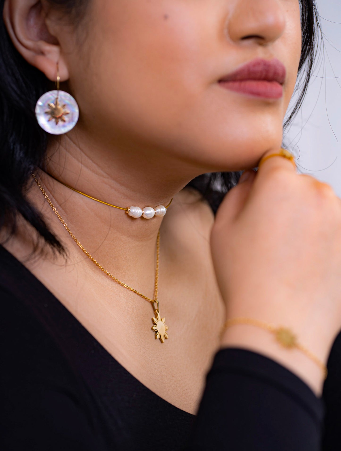 A lifestyle close-up of a person wearing an Aking Sinta jewelry set, featuring the gold Araw necklace layered with a delicate pearl choker. The styling includes matching mother-of-pearl Araw drop earrings and a sun-motif gold bracelet, showcased against a black outfit for an elegant, high-contrast evening look.