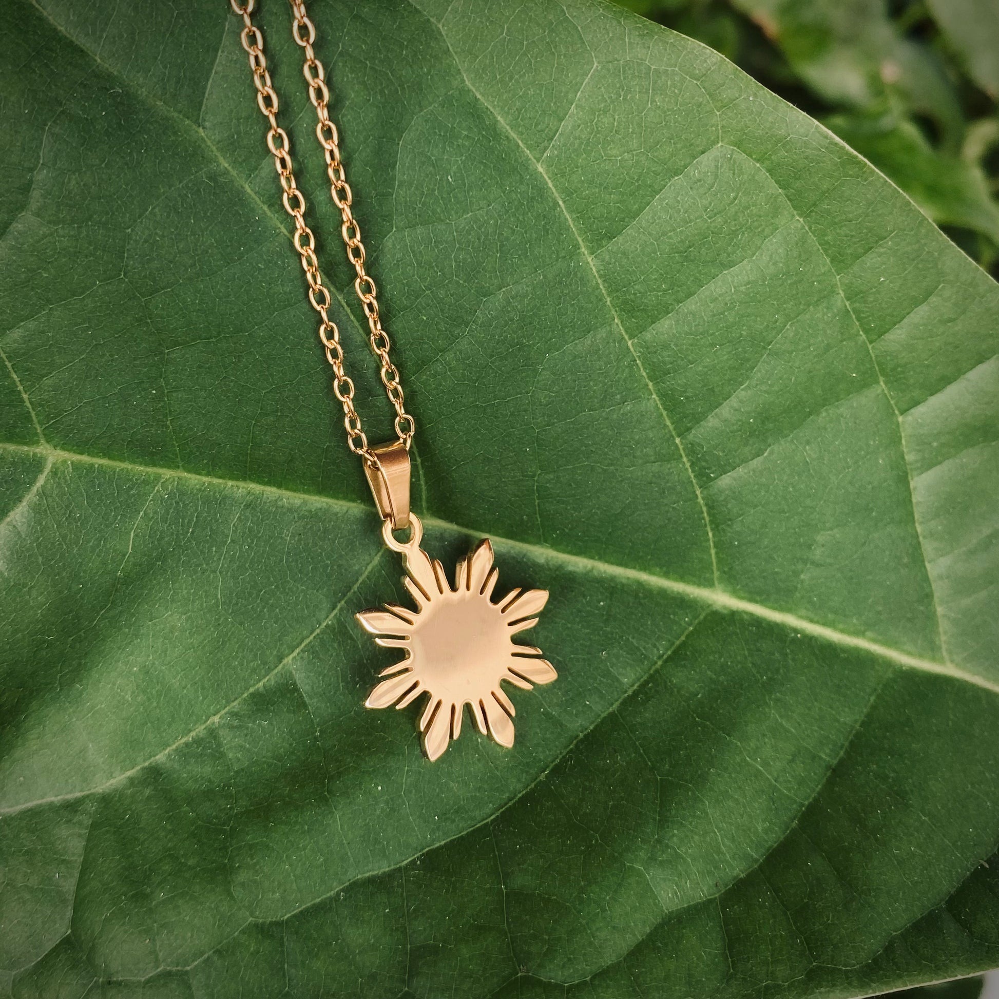 An Aking Sinta minimalist gold necklace featuring a polished eight-rayed "Araw" sun pendant, inspired by the Philippine flag. The pendant rests on a large, vibrant green leaf, creating a natural and organic backdrop that highlights the warm gold tones and clean geometric lines of the Filipino-inspired design.