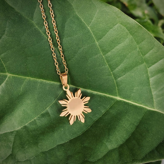 An Aking Sinta minimalist gold necklace featuring a polished eight-rayed "Araw" sun pendant, inspired by the Philippine flag. The pendant rests on a large, vibrant green leaf, creating a natural and organic backdrop that highlights the warm gold tones and clean geometric lines of the Filipino-inspired design.