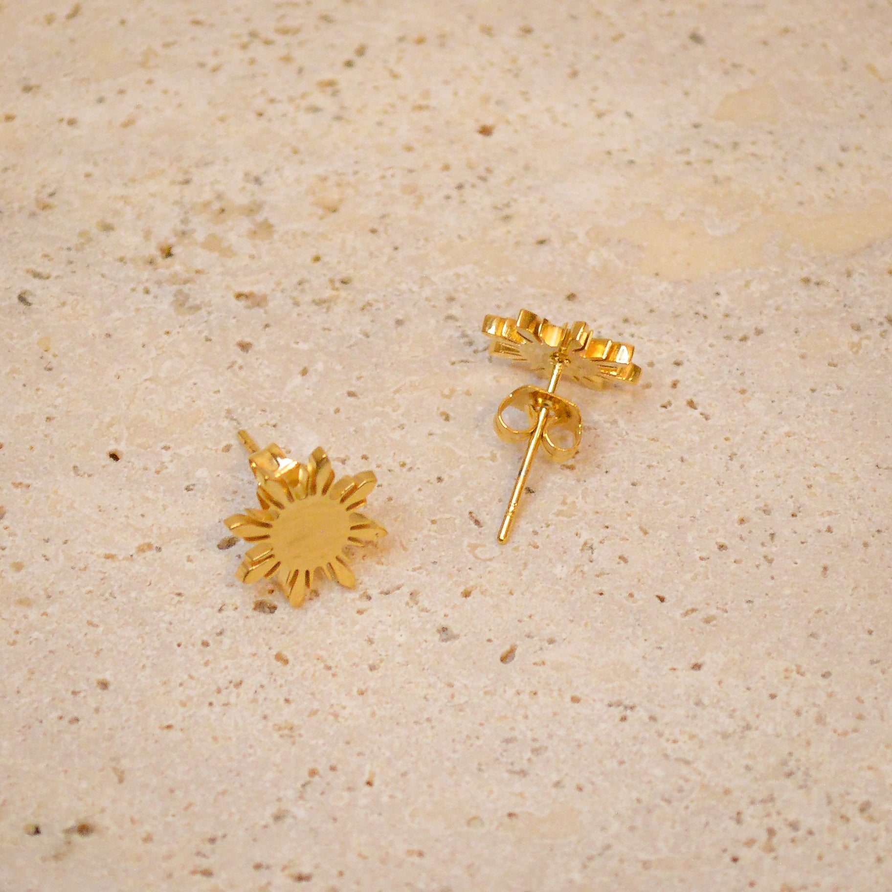 An angled close-up of a pair of Aking Sinta gold Araw stud earrings on a light, textured stone surface. The shot features one earring lying flat and the other tilted to show the butterfly back closure and the thickness of the polished sunburst design, inspired by Filipino heritage