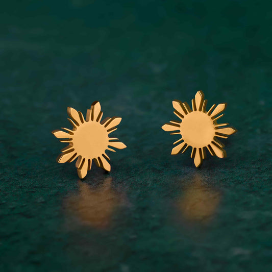 A pair of Aking Sinta gold stud earrings featuring the iconic eight-rayed "Araw" sun from the Philippine flag. The minimalist, polished gold suns are shown standing on a dark teal textured surface with soft lighting reflections, highlighting their sleek and symbolic design