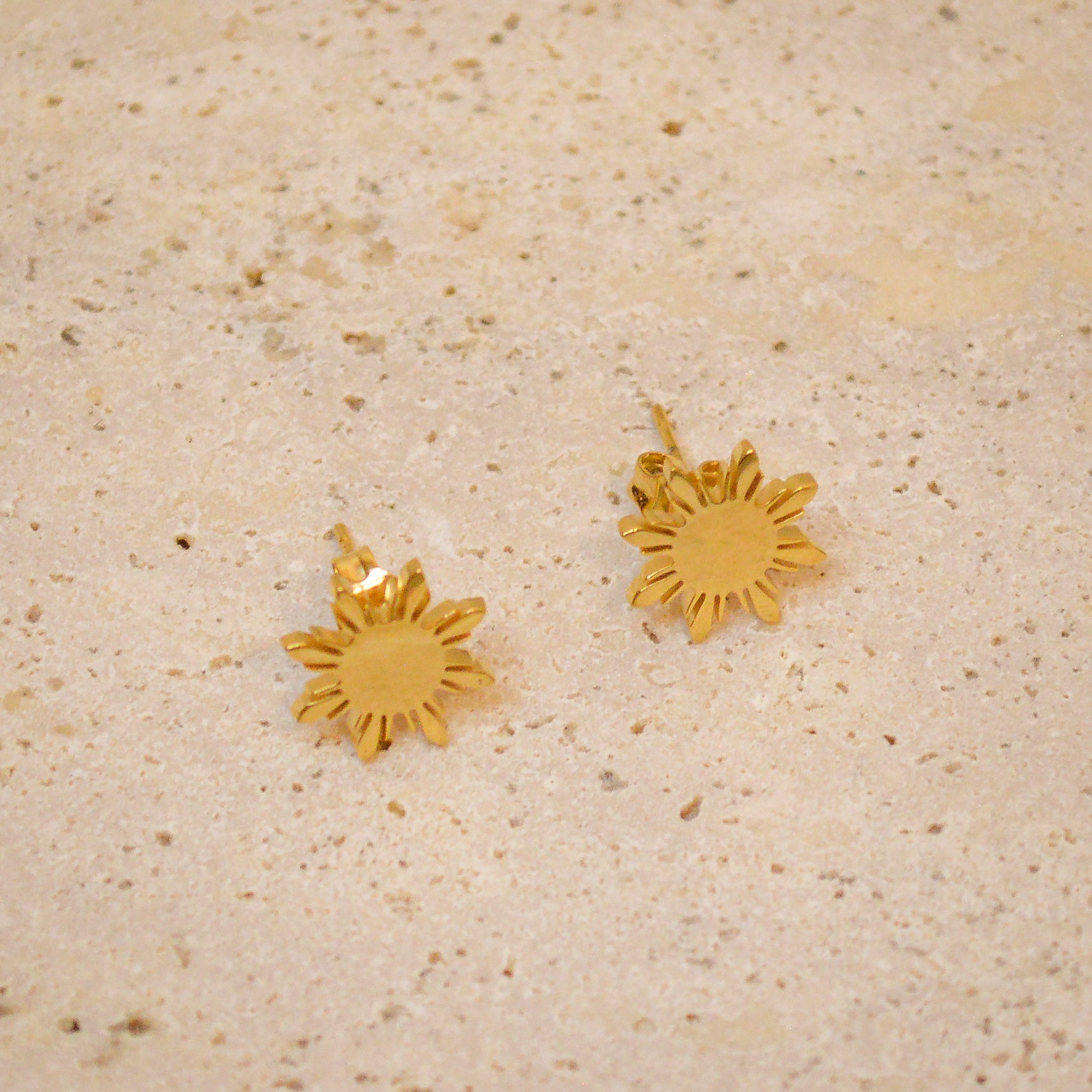 A top-down, high-detail shot of a pair of Aking Sinta gold Araw stud earrings. The polished gold finish highlights the eight-rayed Filipino sun design, beautifully contrasted against the neutral tones of a textured natural stone surface.