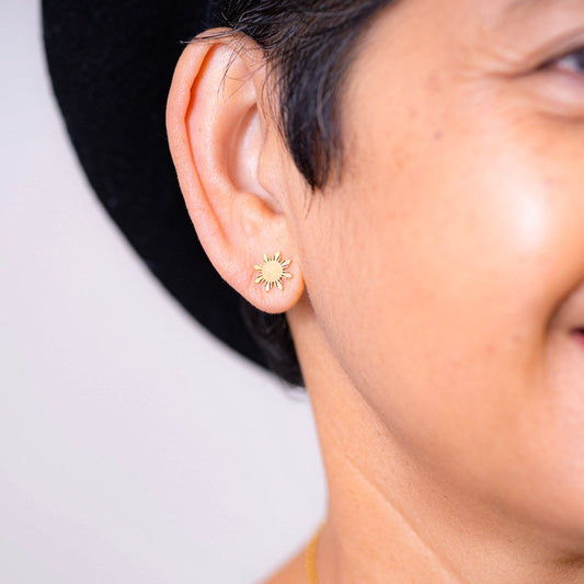 A lifestyle close-up of a person wearing an Aking Sinta gold Araw stud earring. The minimalist eight-rayed sun design sits elegantly on the earlobe, showcasing its delicate scale and polished finish as part of a modern Filipino-inspired jewelry look