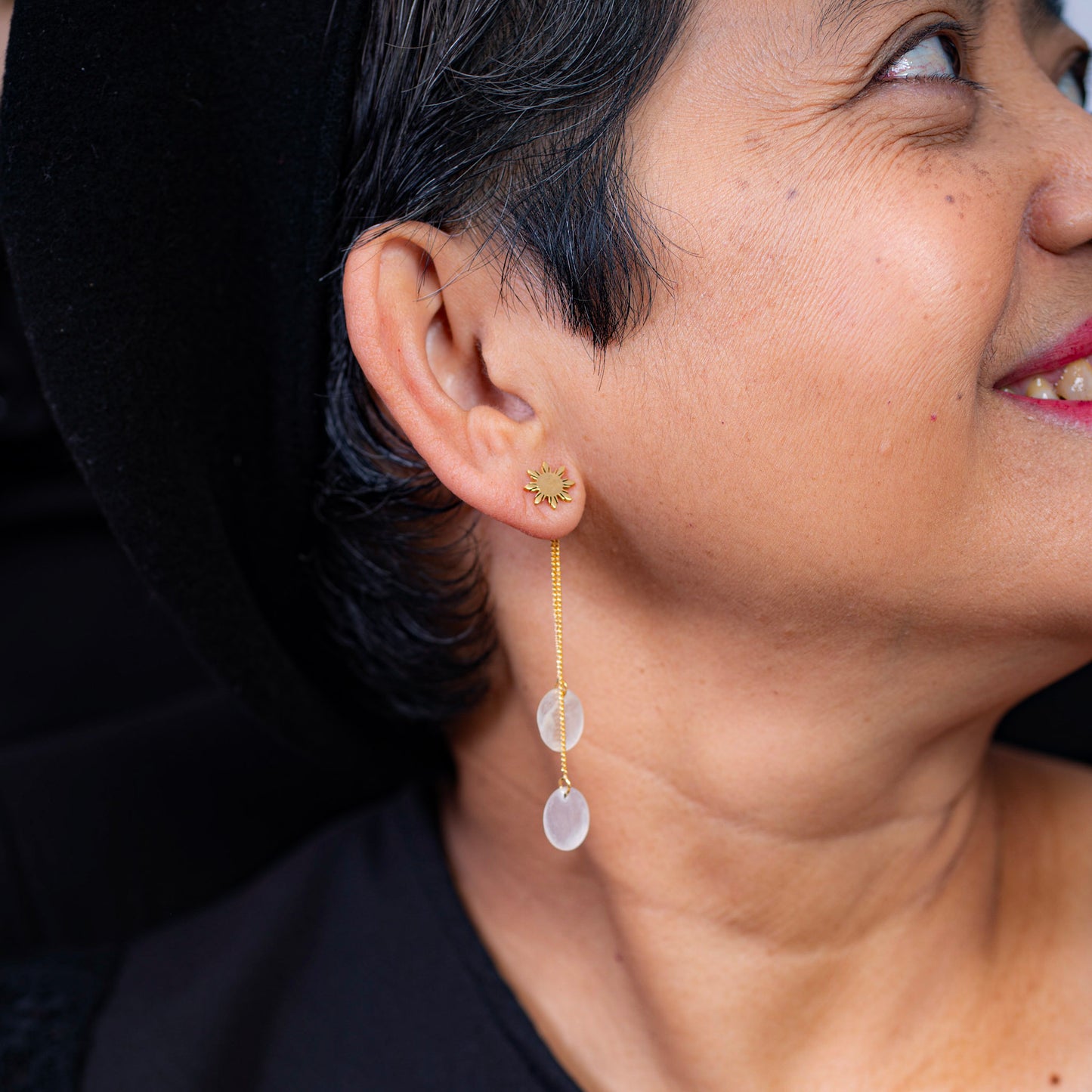 A lifestyle close-up of a person wearing an Aking Sinta gold Araw stud earring paired with a long, delicate gold chain drop featuring two translucent white mother-of-pearl discs. The minimalist eight-rayed sun design is shown in a sophisticated styling context against a dark background, highlighting the versatility of the Filipino-inspired piece.
