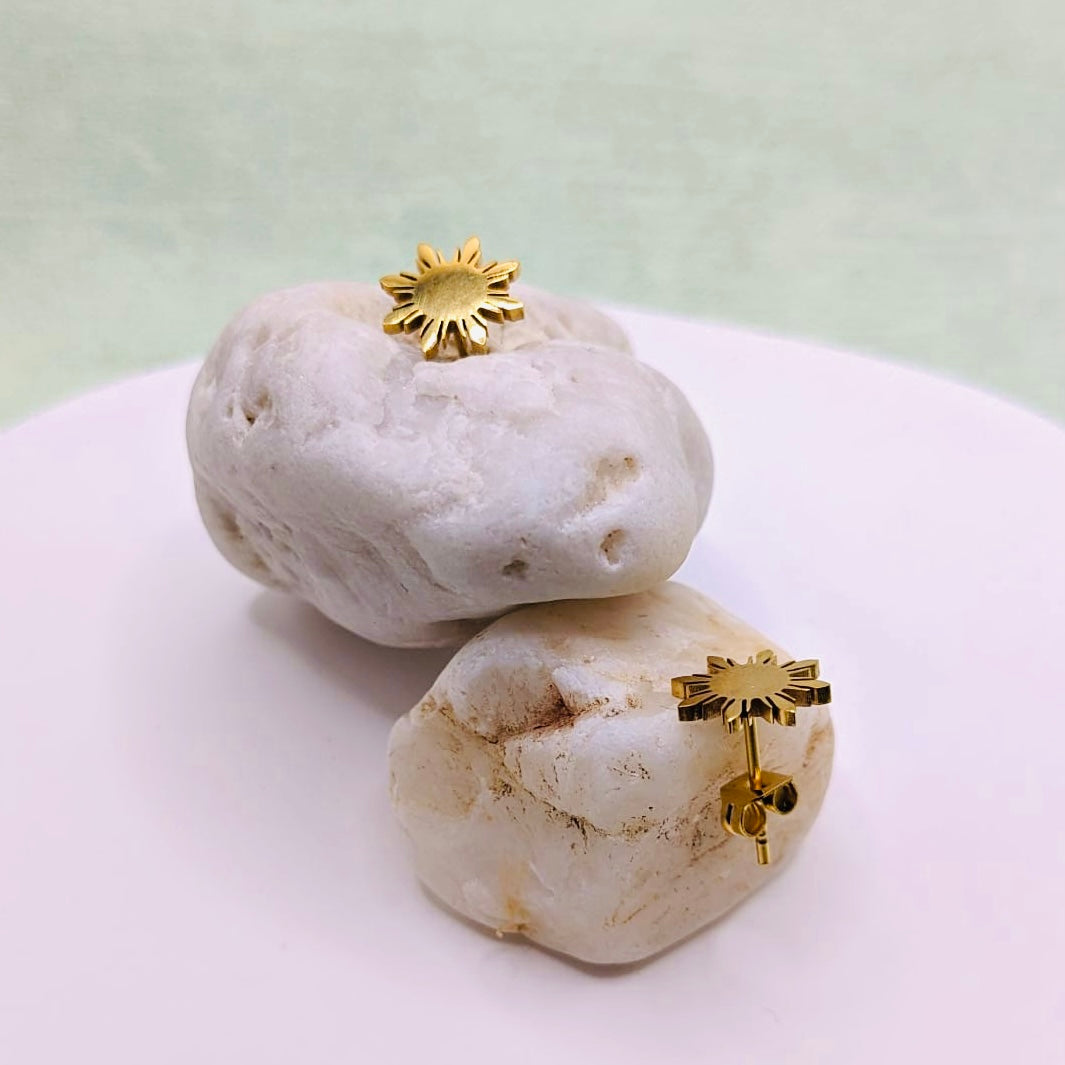 A pair of Aking Sinta gold Araw stud earrings artistically displayed on stacked white river stones. The minimalist eight-rayed sun design, inspired by the Philippine flag, is shown from both a top and side profile to highlight the polished finish and sturdy butterfly back closures against a clean, light background