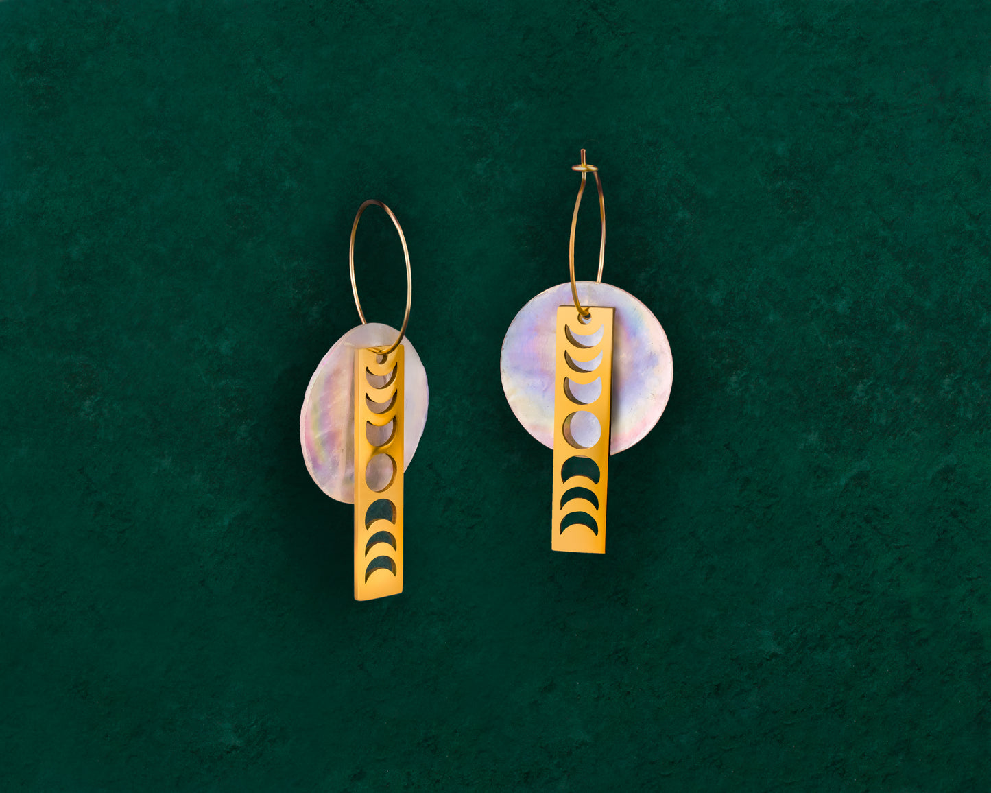 A pair of Aking Sinta gold bar earrings on a dark green background; each long, rectangular gold bar features a vertical series of moon phase cutouts (crescent to full moon) with a small, round white Capiz shell charm dangling from the bottom.