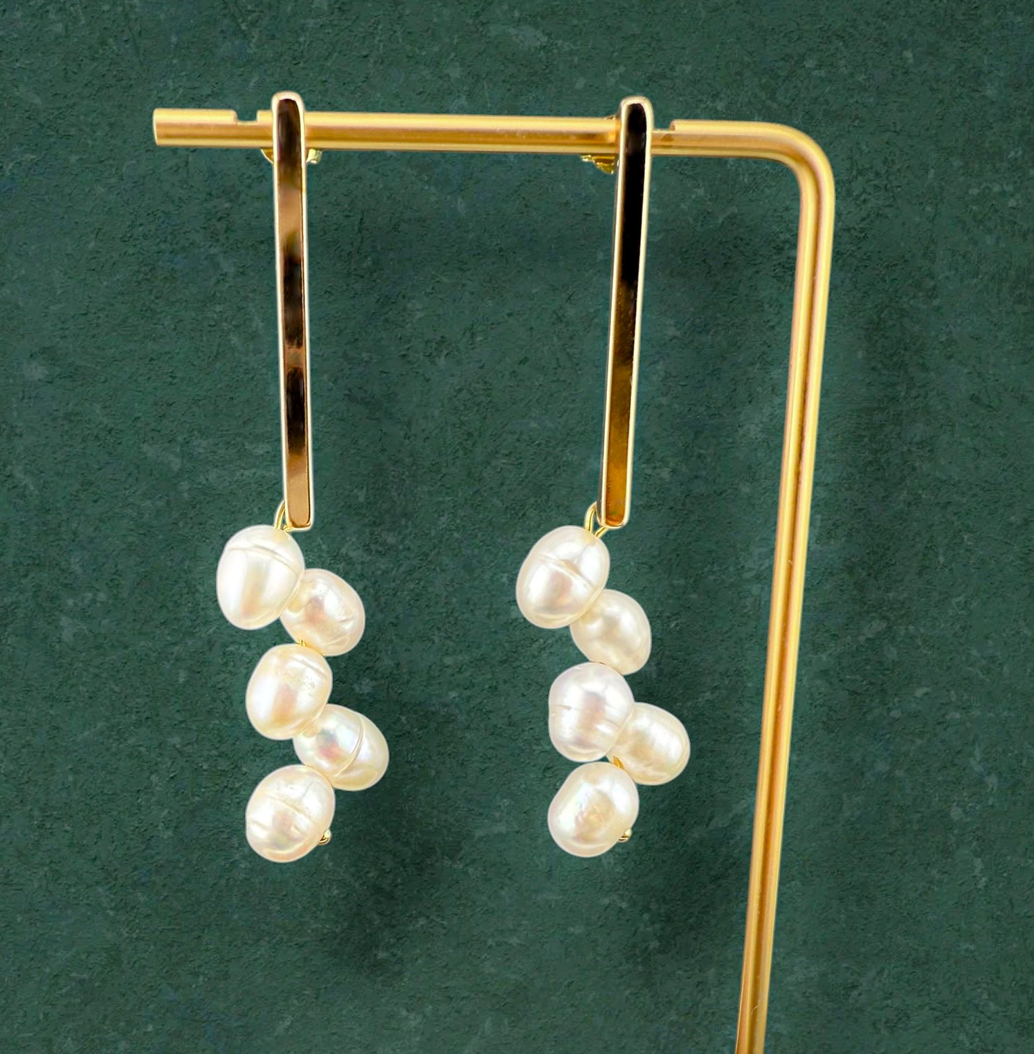 A pair of Aking Sinta gold bar stud earrings with white freshwater pearl clusters hanging from them. The earrings are showcased on a gold T-bar jewelry stand against a dark green textured background