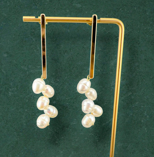 A pair of Aking Sinta gold bar stud earrings with white freshwater pearl clusters hanging from them. The earrings are showcased on a gold T-bar jewelry stand against a dark green textured background