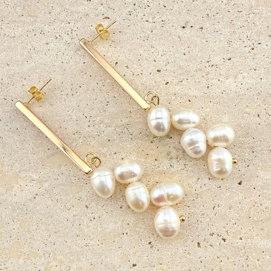 A pair of Aking Sinta gold bar stud earrings featuring a dangling "wheat" cluster of white freshwater pearls. The modern, elegant design combines a polished gold horizontal bar with organic, lustrous pearl drops, displayed on a light natural stone surface.