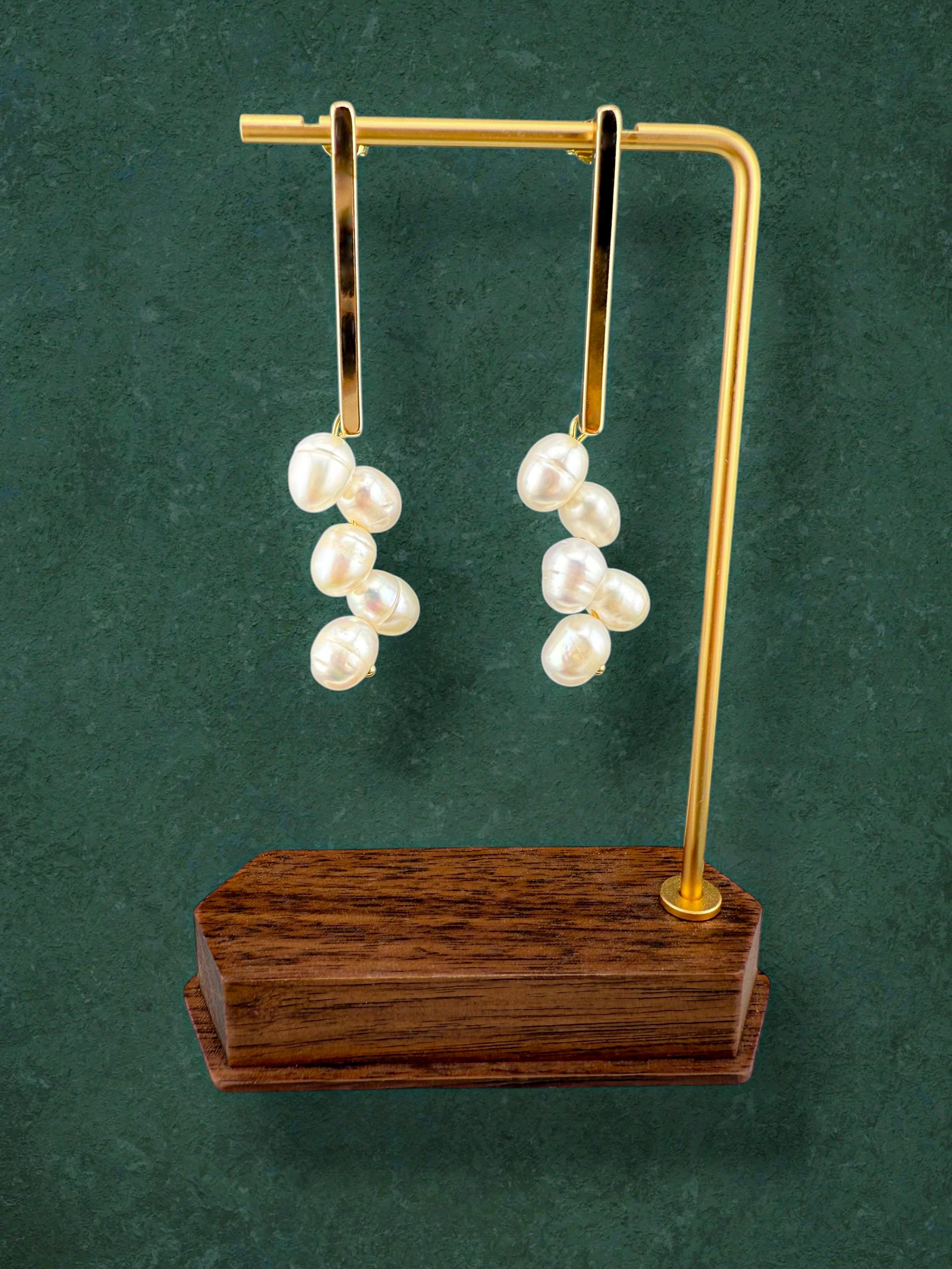A pair of Aking Sinta gold bar stud earrings with white freshwater pearl clusters, displayed on a gold T-bar stand with a dark wood base. The elegant jewelry is set against a dark green textured background, highlighting the lustrous white pearls