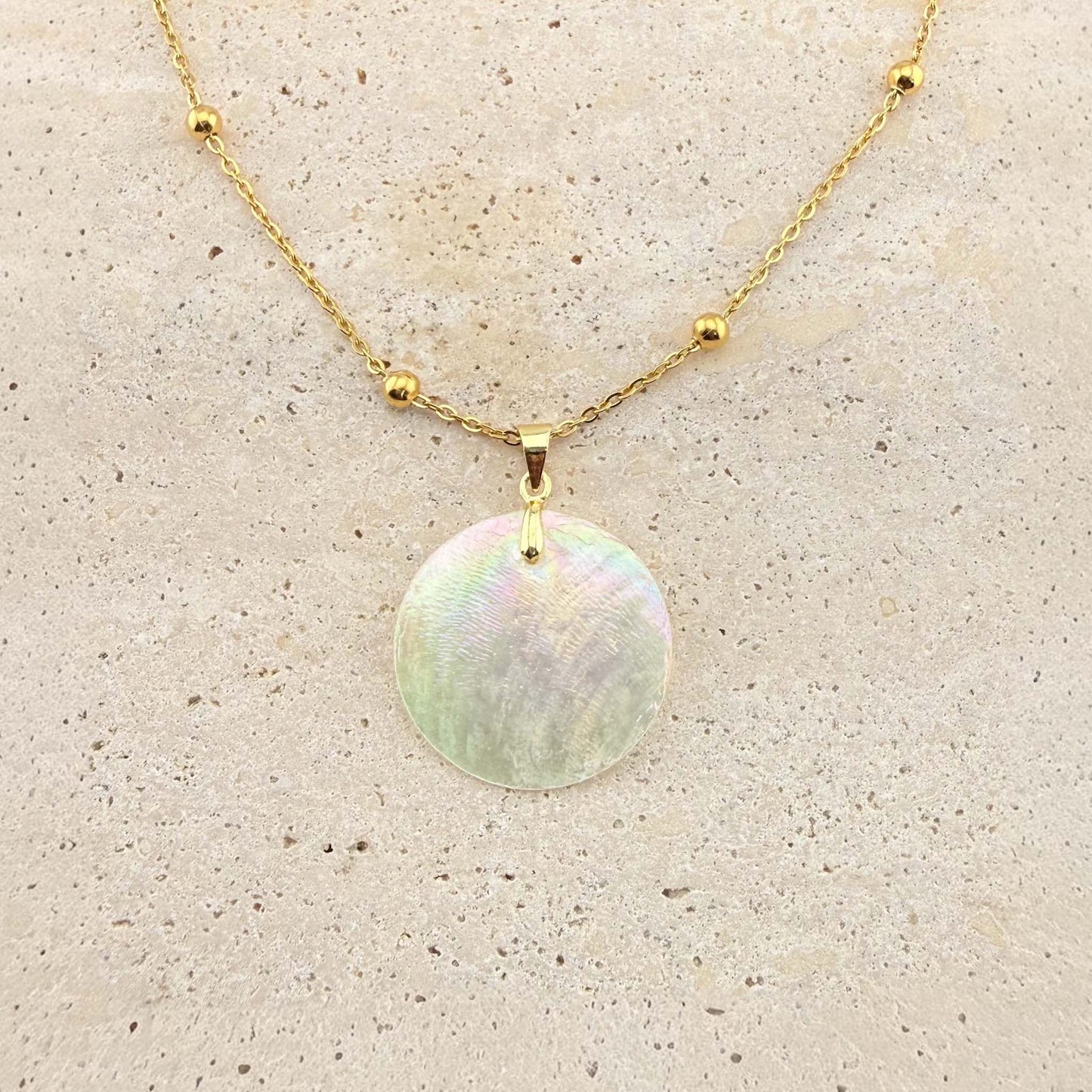 A gold Aking Sinta necklace featuring a large, circular, iridescent Capiz shell pendant. The shimmering, natural shell disc is suspended from a polished gold bail on a gold cable chain adorned with small, evenly spaced gold beads. The necklace is displayed on a light, textured natural stone surface