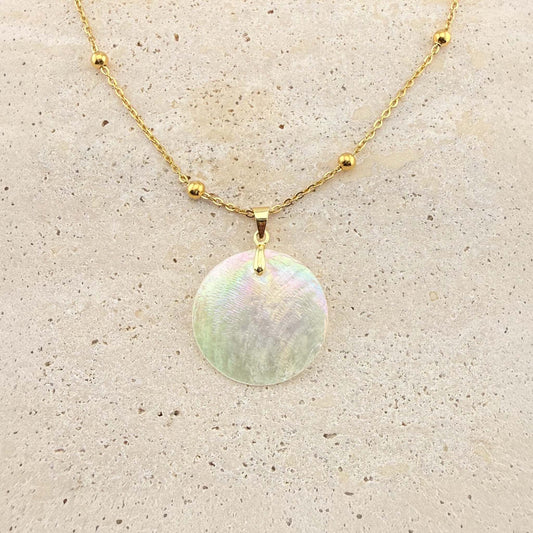 A gold Aking Sinta necklace featuring a large, circular, iridescent Capiz shell pendant. The shimmering, natural shell disc is suspended from a polished gold bail on a gold cable chain adorned with small, evenly spaced gold beads. The necklace is displayed on a light, textured natural stone surface
