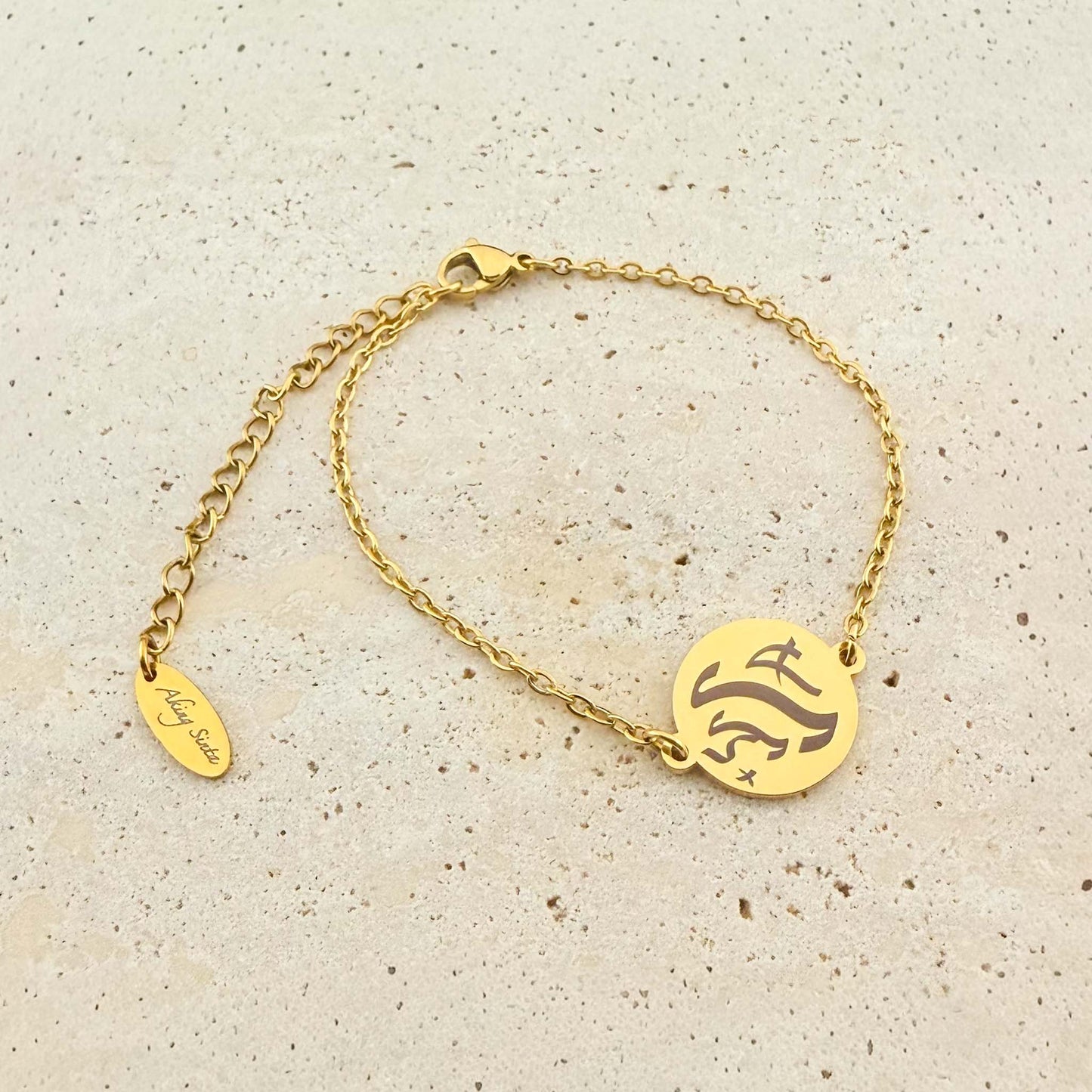 A gold Aking Sinta coin bracelet featuring a bespoke engraving in ancient Filipino Baybayin script. The bracelet includes a lobster clasp and an adjustable extender chain with a signature "Aking Sinta" oval tag, displayed on a light natural stone surface.