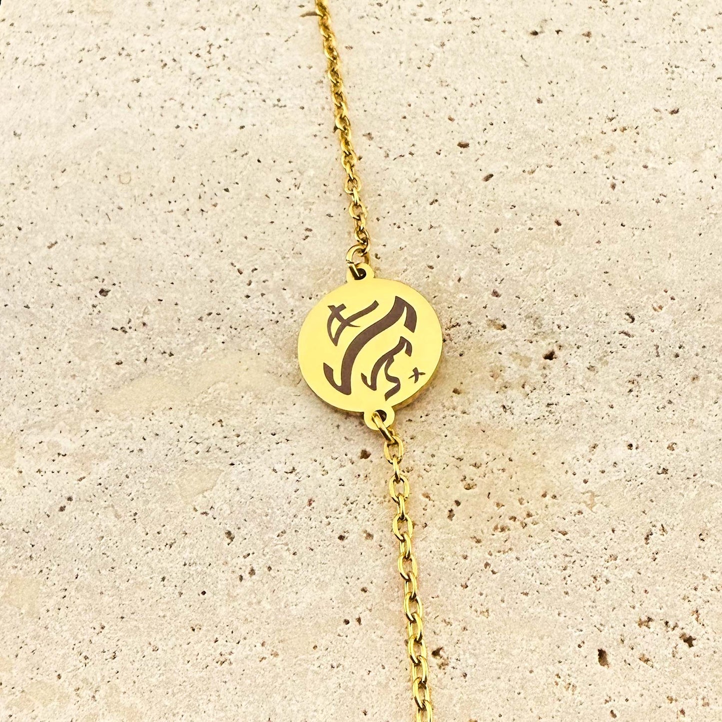 A close-up of a gold Aking Sinta coin bracelet featuring a bespoke engraving in ancient Filipino Baybayin script. The circular gold pendant is centered on a delicate link chain and displayed on a light, textured natural stone surface