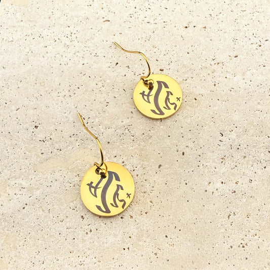 A pair of gold Aking Sinta hook earrings featuring circular coin charms with bespoke engravings. The charms are etched with ancient Filipino Baybayin script and hang from elegant gold fishhook wires, displayed on a light-colored natural stone surface