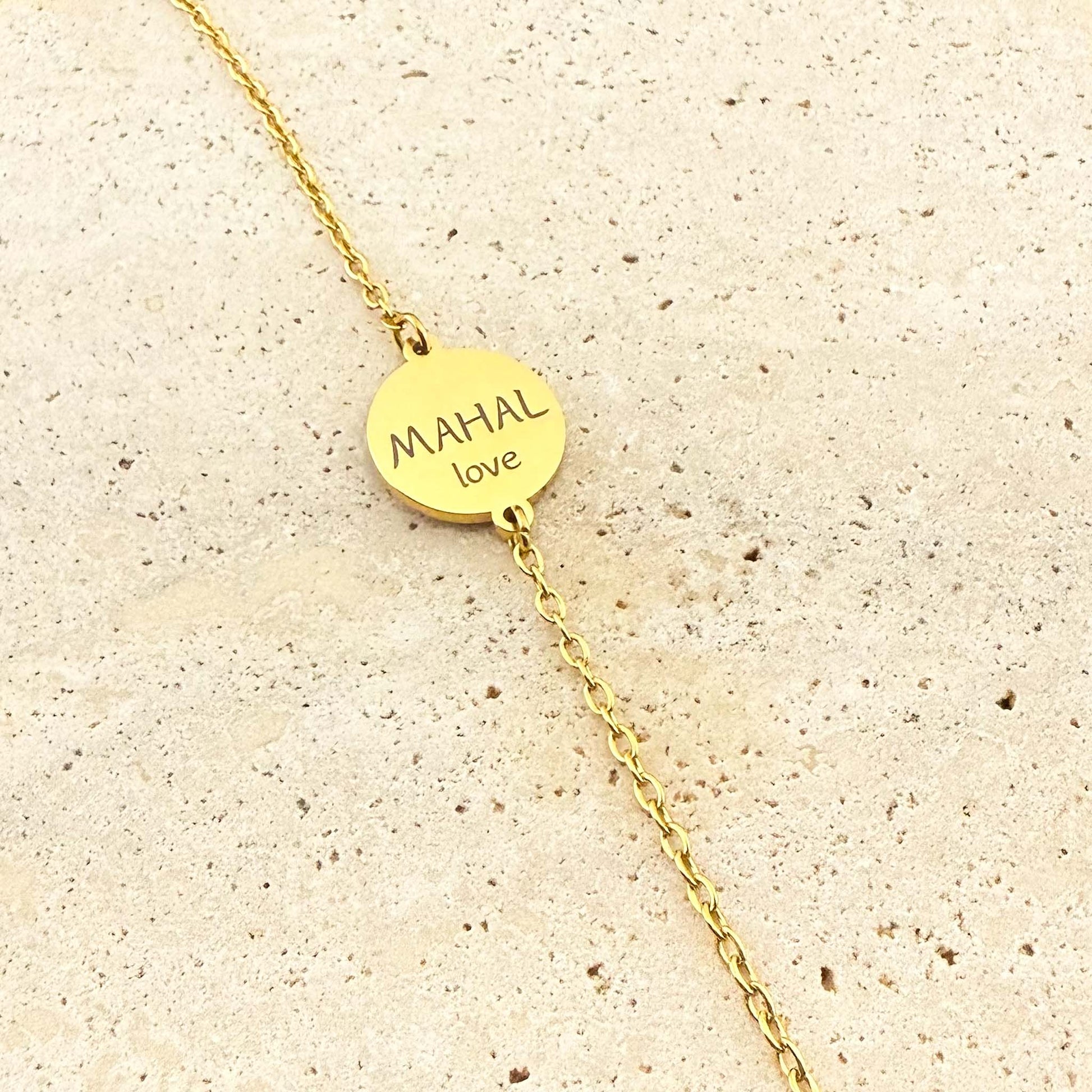 A gold Aking Sinta coin bracelet featuring a bespoke engraving. The circular gold pendant is engraved with the word "MAHAL" and its English translation "love" in a clean sans-serif font. The jewelry is centered on a light-colored natural stone surface.
