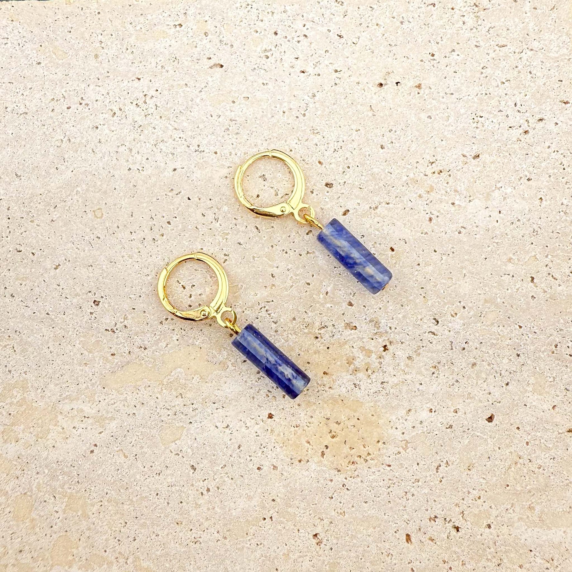 Gold Aking Sinta huggie earrings with rectangular blue watermelon column-cut glass charms on a white marble surface.

