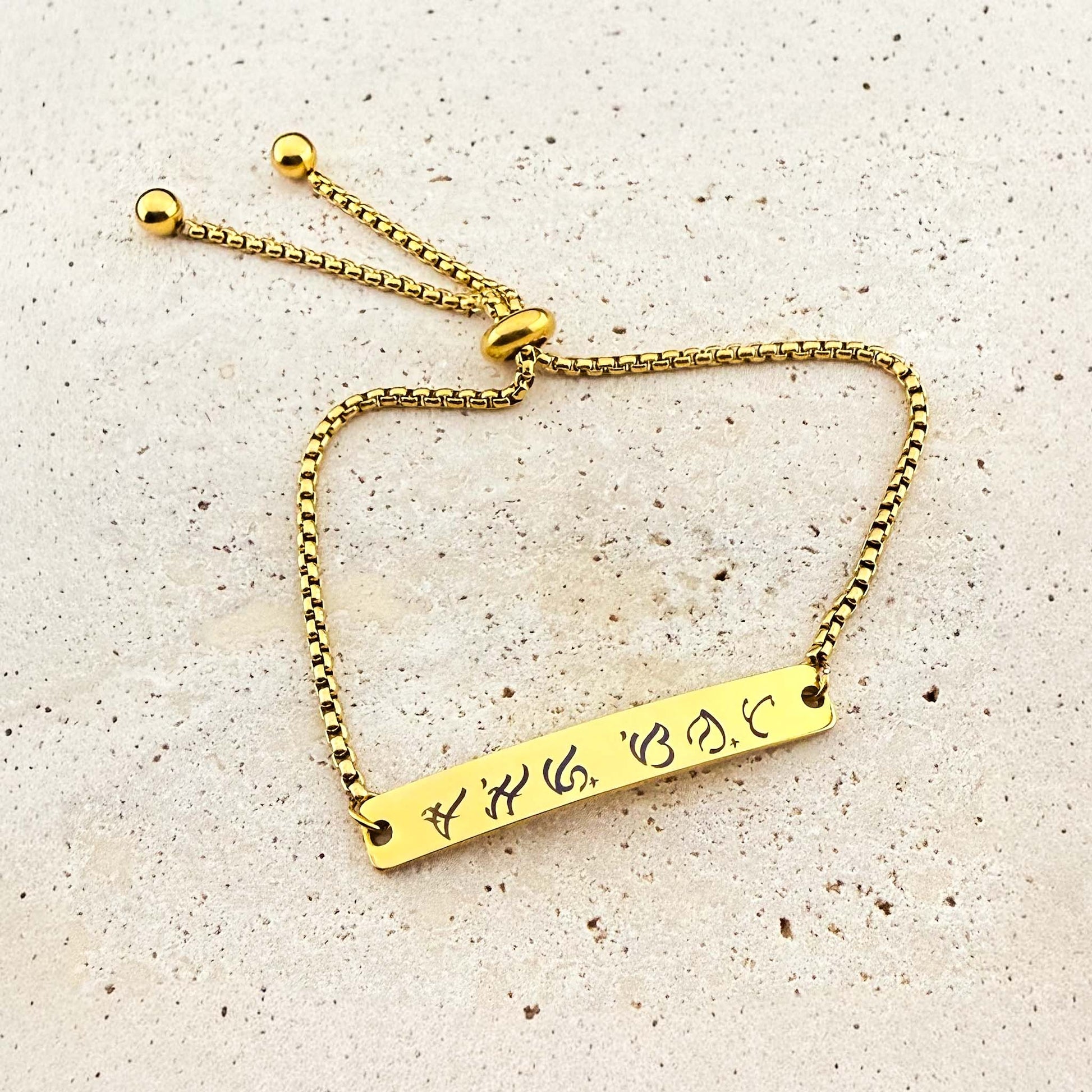 A gold Aking Sinta box chain bracelet featuring a bespoke engraving. The flat gold bar is etched with ancient Filipino Baybayin script. The jewelry includes an adjustable bolo-style slider clasp with spherical end beads, displayed on a light natural stone surface