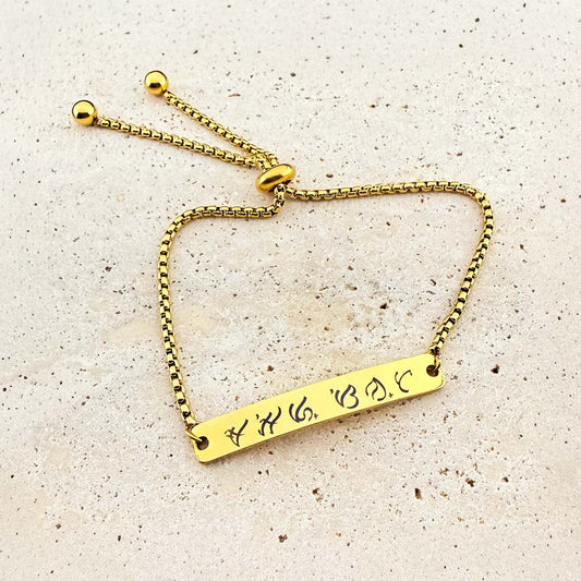 A gold Aking Sinta box chain bracelet featuring a bespoke engraving. The flat gold bar is etched with ancient Filipino Baybayin script. The jewelry includes an adjustable bolo-style slider clasp with spherical end beads, displayed on a light natural stone surface