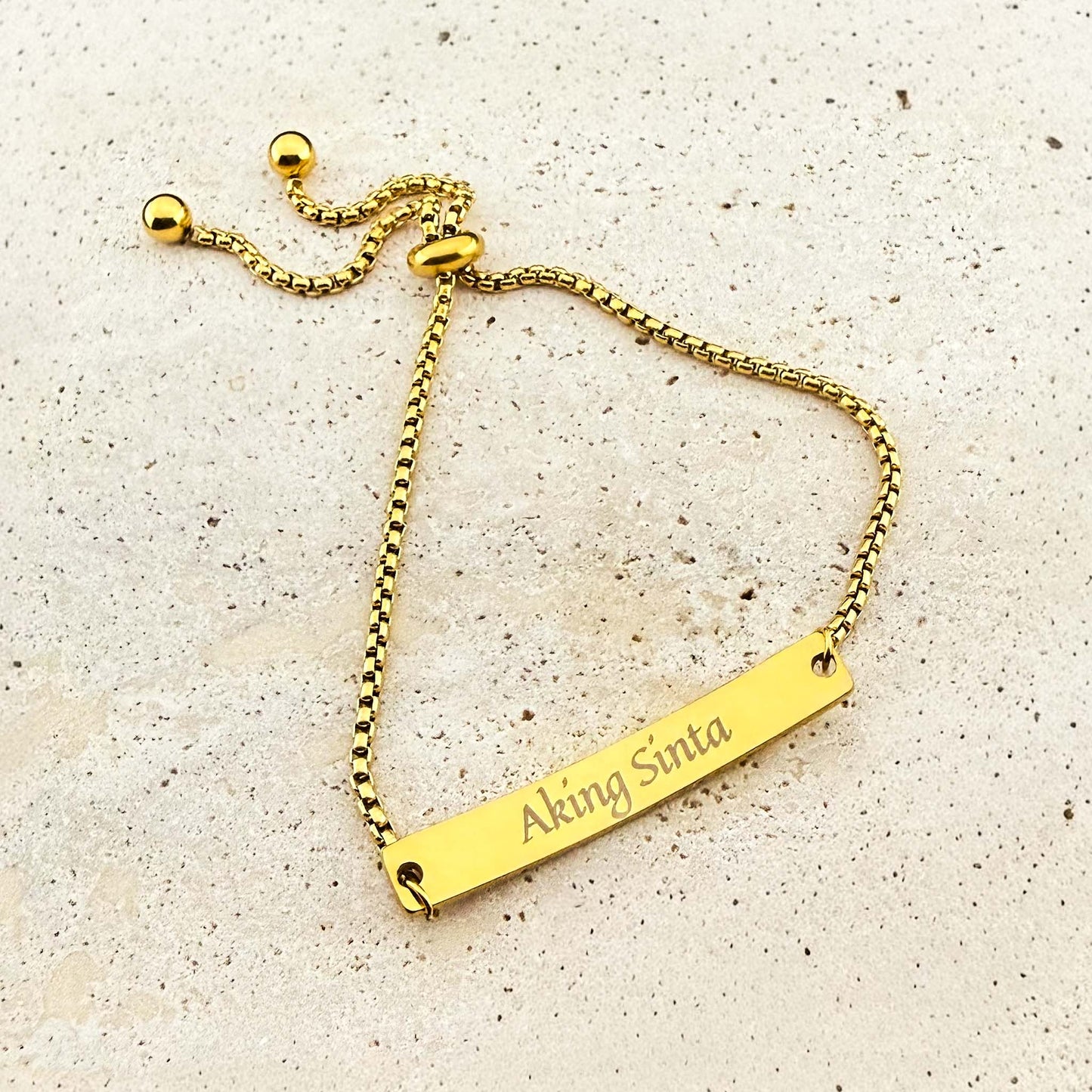A gold Aking Sinta box chain bracelet featuring a bespoke engraving. The flat gold bar is etched with the "Aking Sinta" brand logo in a cursive font. The jewelry includes an adjustable bolo-style slider clasp with spherical end beads, displayed on a light natural stone surface