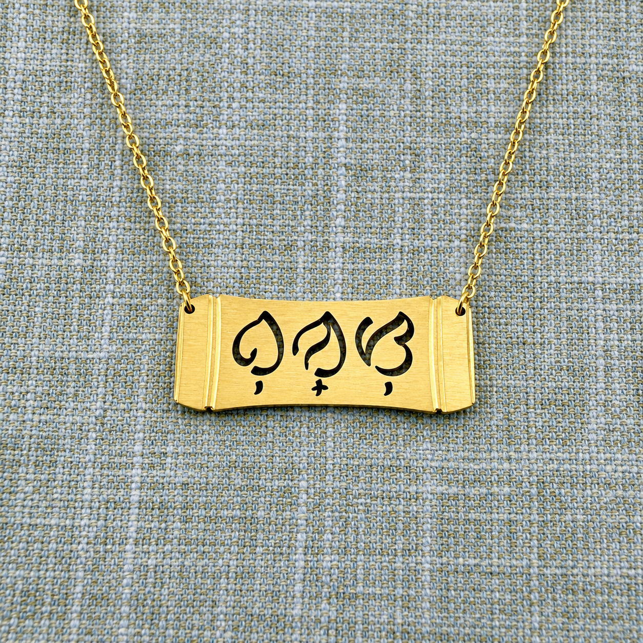 An Aking Sinta gold horizontal bar necklace centered on a textured blue-gray and gold woven linen backdrop. The matte gold pendant features a stylized bamboo section frame with the word "Bunso" (Youngest Child) cleanly cut out in traditional script-like Baybayin characters, suspended from a fine gold link chain