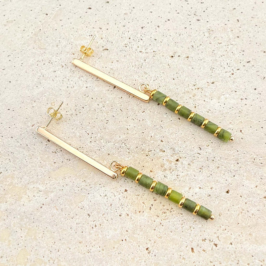 A pair of elegant Aking Sinta gold bar drop stud earrings. Each earring features a sleek, polished gold bar stud from which a long drop hangs, composed of alternating small, round Canadian jade beads and gold spacers. The earrings are displayed on a light-colored natural stone surface