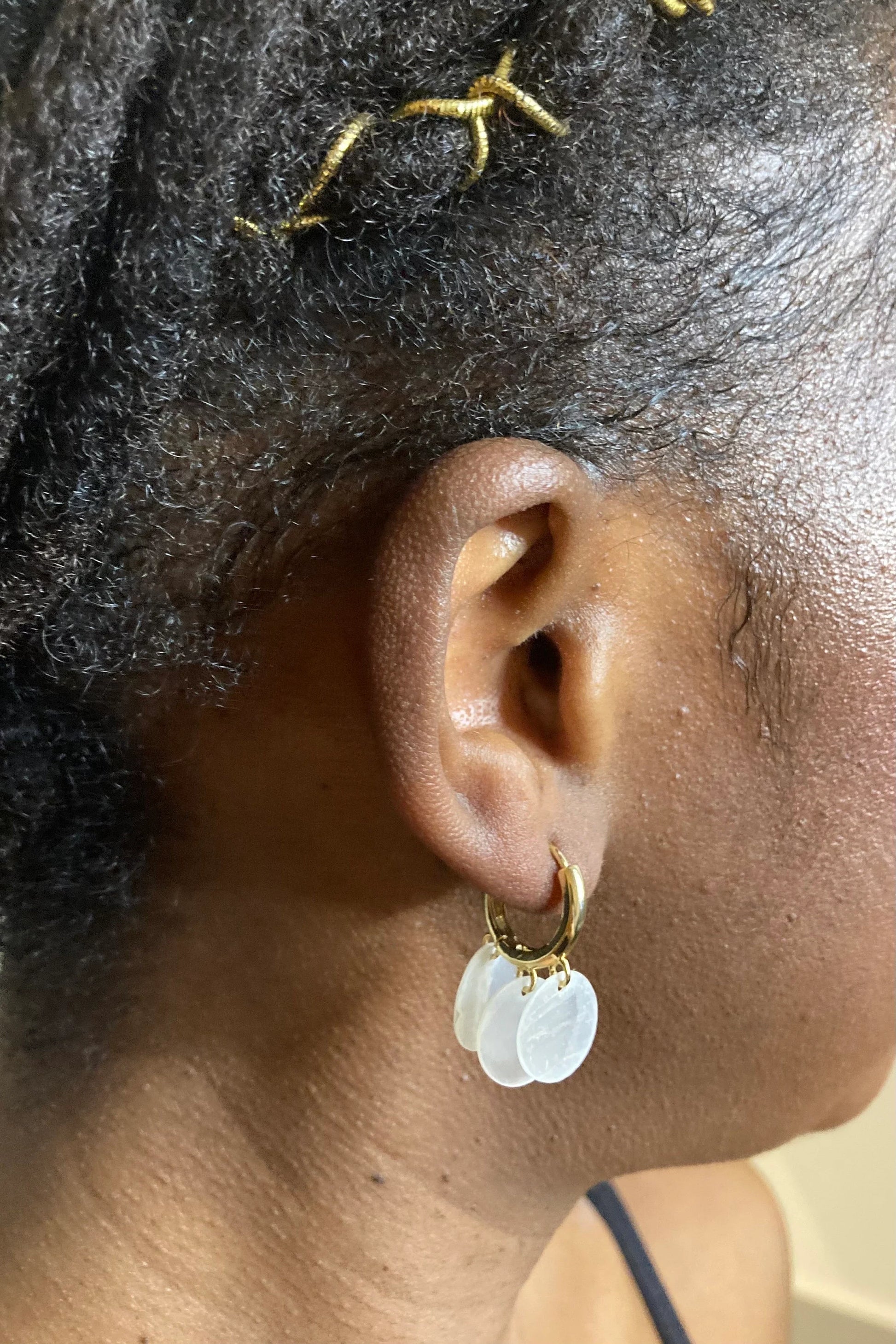 A detailed close-up shot of a model wearing Aking Sinta gold huggie hoop earrings. Each polished gold hoop features three delicate, circular, iridescent Capiz shell charms that catch the light. The model has dark, textured hair styled with gold wire, and the earrings are positioned elegantly on the ear, highlighting their shimmering, artisanal design