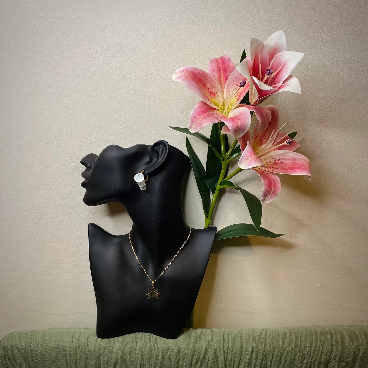 A stylized display of Aking Sinta jewelry on a black mannequin bust. The mannequin wears Aking Sinta gold huggie hoop earrings with dangling, circular Capiz shell charms and a gold sun-motif pendant necklace. The display is elegantly complemented by a vibrant pink lily flower, set against a simple, warm-toned wall
