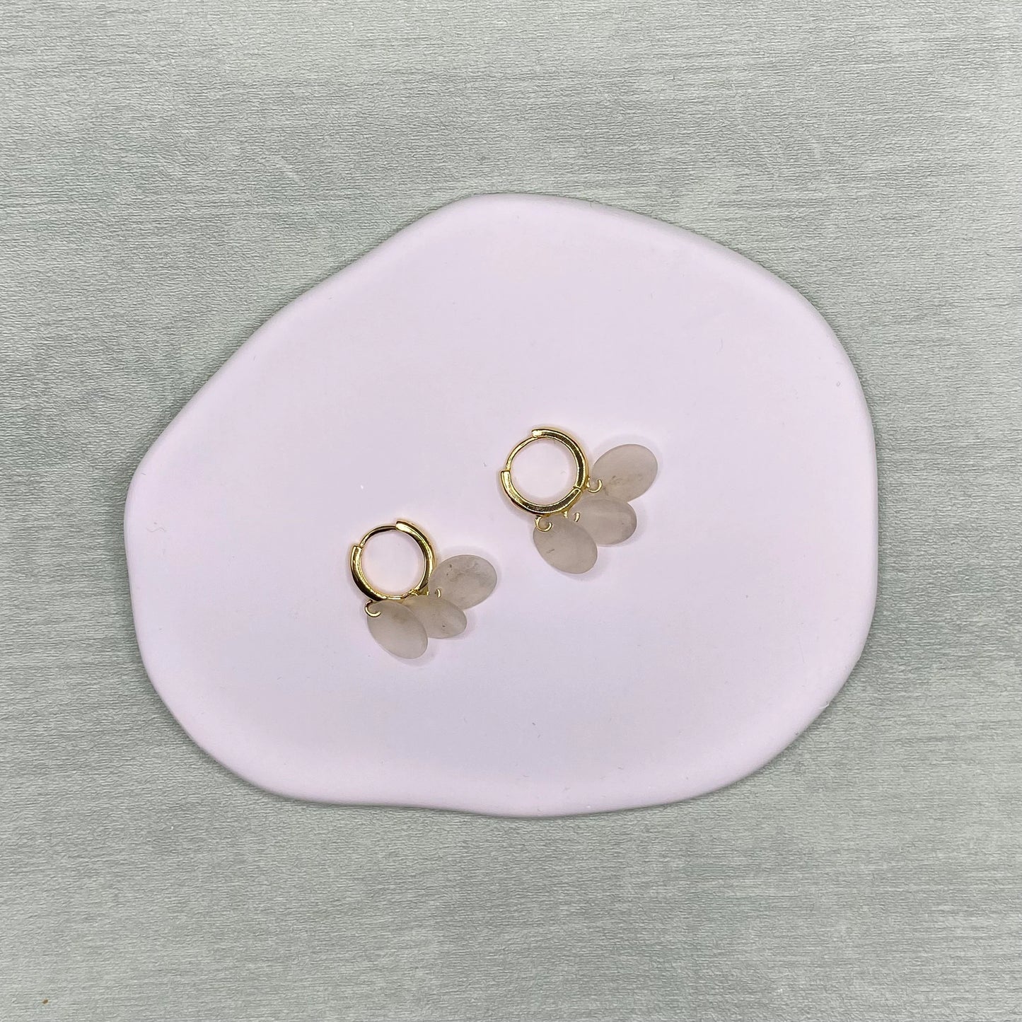 A flat-lay photograph of a pair of Aking Sinta gold huggie hoop earrings, each featuring three small, dangling circular Capiz shell charms. The delicate earrings are centered on a smooth, organic-shaped light pink jewelry tray, set against a neutral, textured grey surface