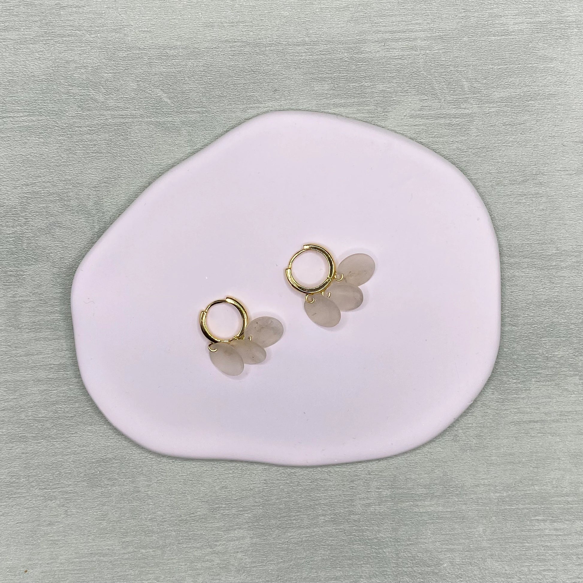A flat-lay photograph of a pair of Aking Sinta gold huggie hoop earrings, each featuring three small, dangling circular Capiz shell charms. The delicate earrings are centered on a smooth, organic-shaped light pink jewelry tray, set against a neutral, textured grey surface