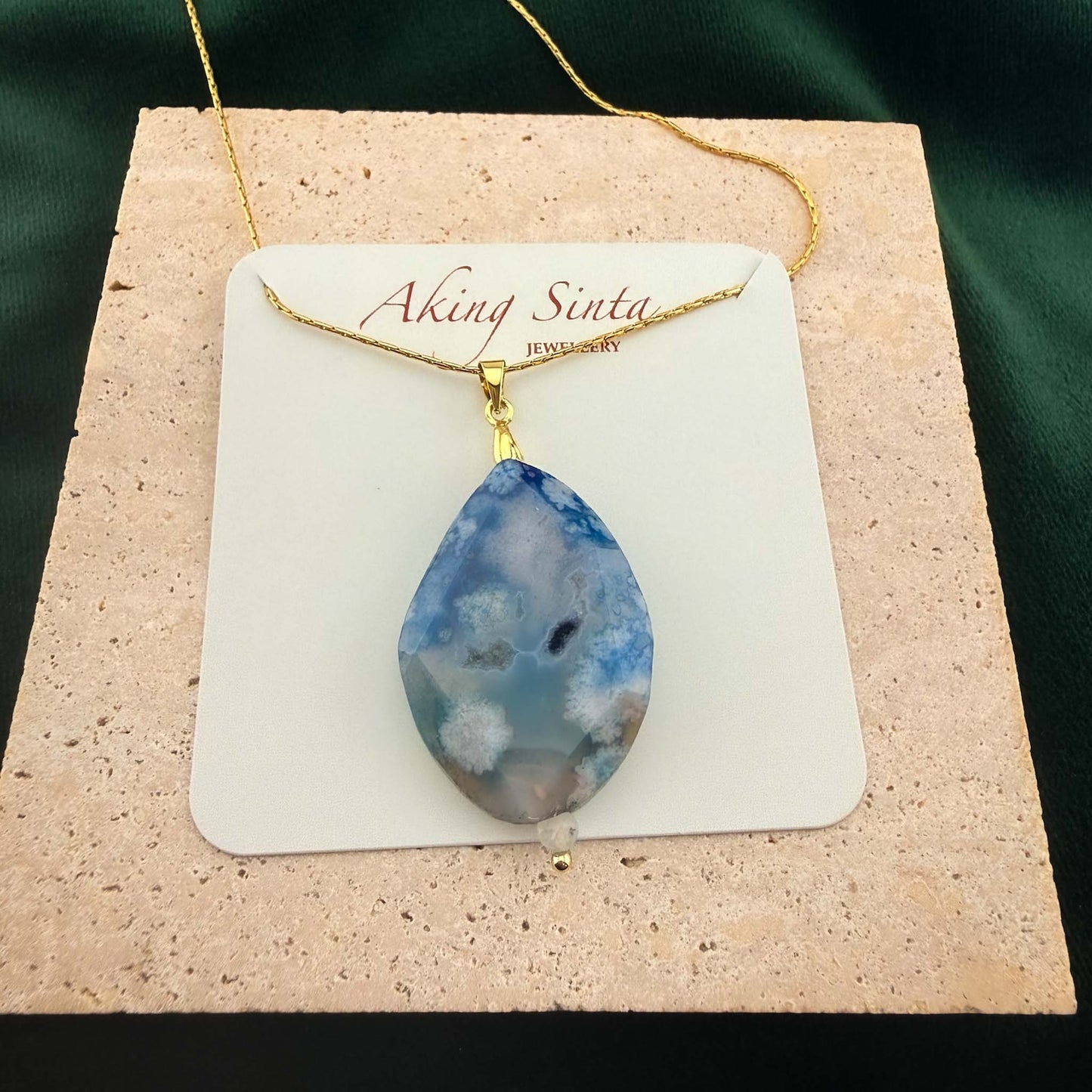 A beautifully packaged Aking Sinta gold chain necklace featuring a faceted teardrop blue agate pendant, displayed on a branded white jewelry card. The necklace rests on a light, textured natural stone block set against a rich dark green background, highlighting both the branding and the unique gemstone quality