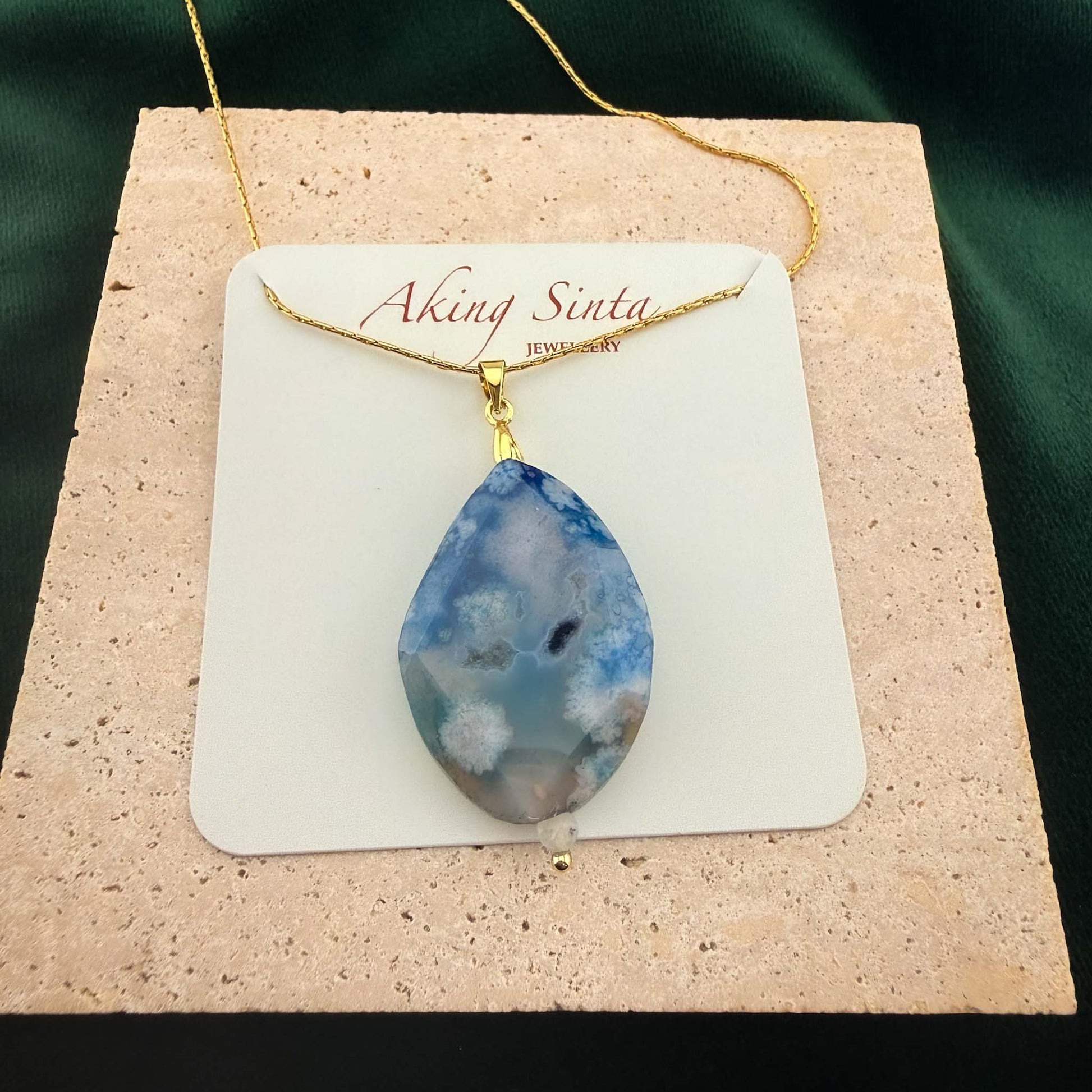 A beautifully packaged Aking Sinta gold chain necklace featuring a faceted teardrop blue agate pendant, displayed on a branded white jewelry card. The necklace rests on a light, textured natural stone block set against a rich dark green background, highlighting both the branding and the unique gemstone quality