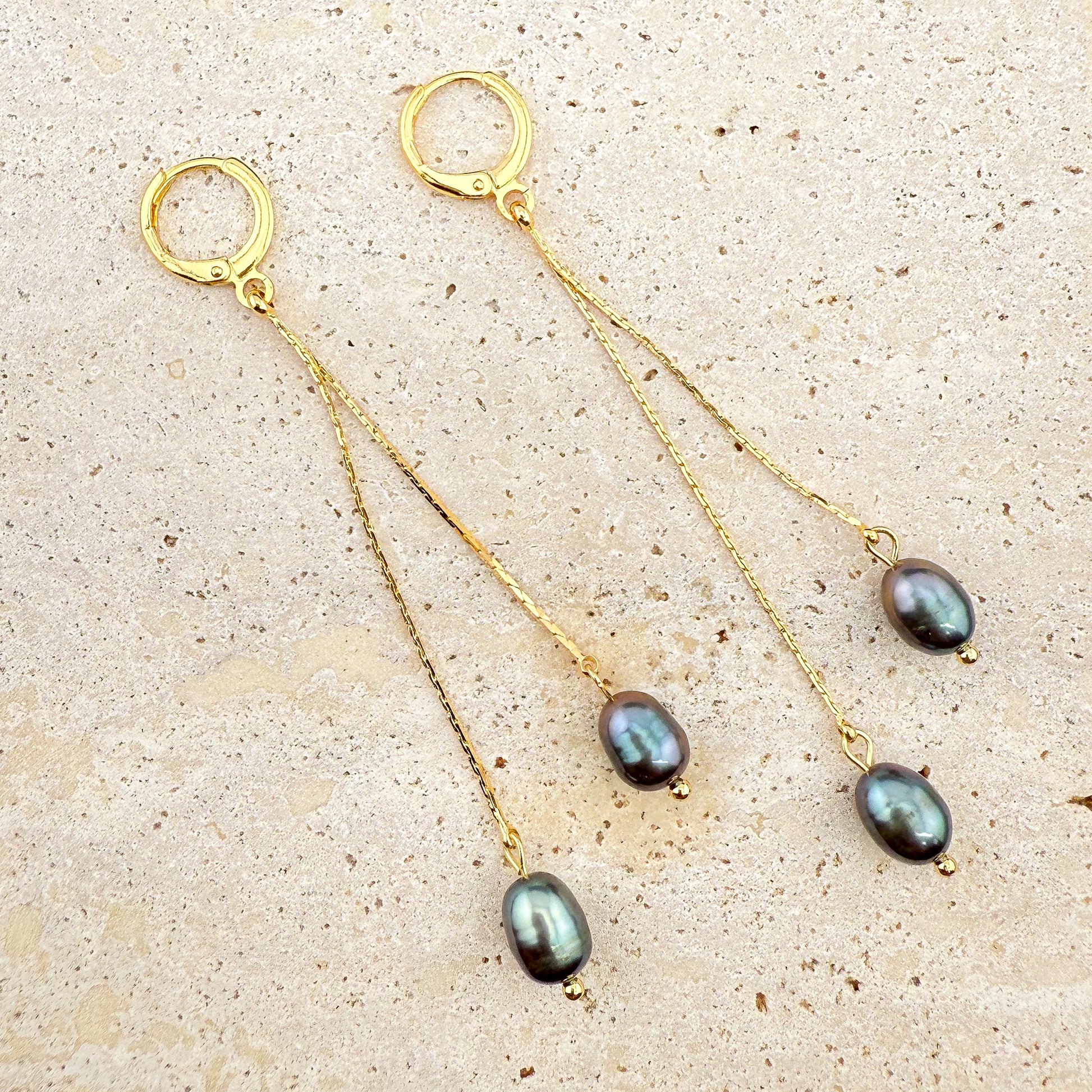 A pair of elegant Aking Sinta gold huggie hoop earrings, each featuring two delicate, dangling gold chains of varying lengths, finished with lustrous dark, iridescent grey-tone pearls. The earrings are arranged on a light, textured natural stone background, emphasizing the contrast between the polished gold and the deep, moody pearls