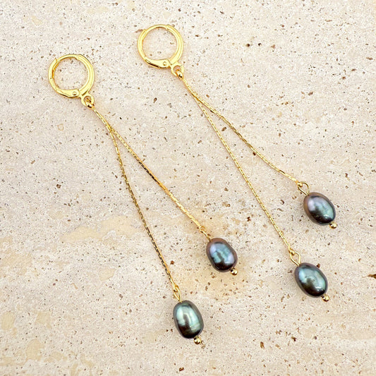 A pair of elegant Aking Sinta gold huggie hoop earrings, each featuring two delicate, dangling gold chains of varying lengths, finished with lustrous dark, iridescent grey-tone pearls. The earrings are arranged on a light, textured natural stone background, emphasizing the contrast between the polished gold and the deep, moody pearls