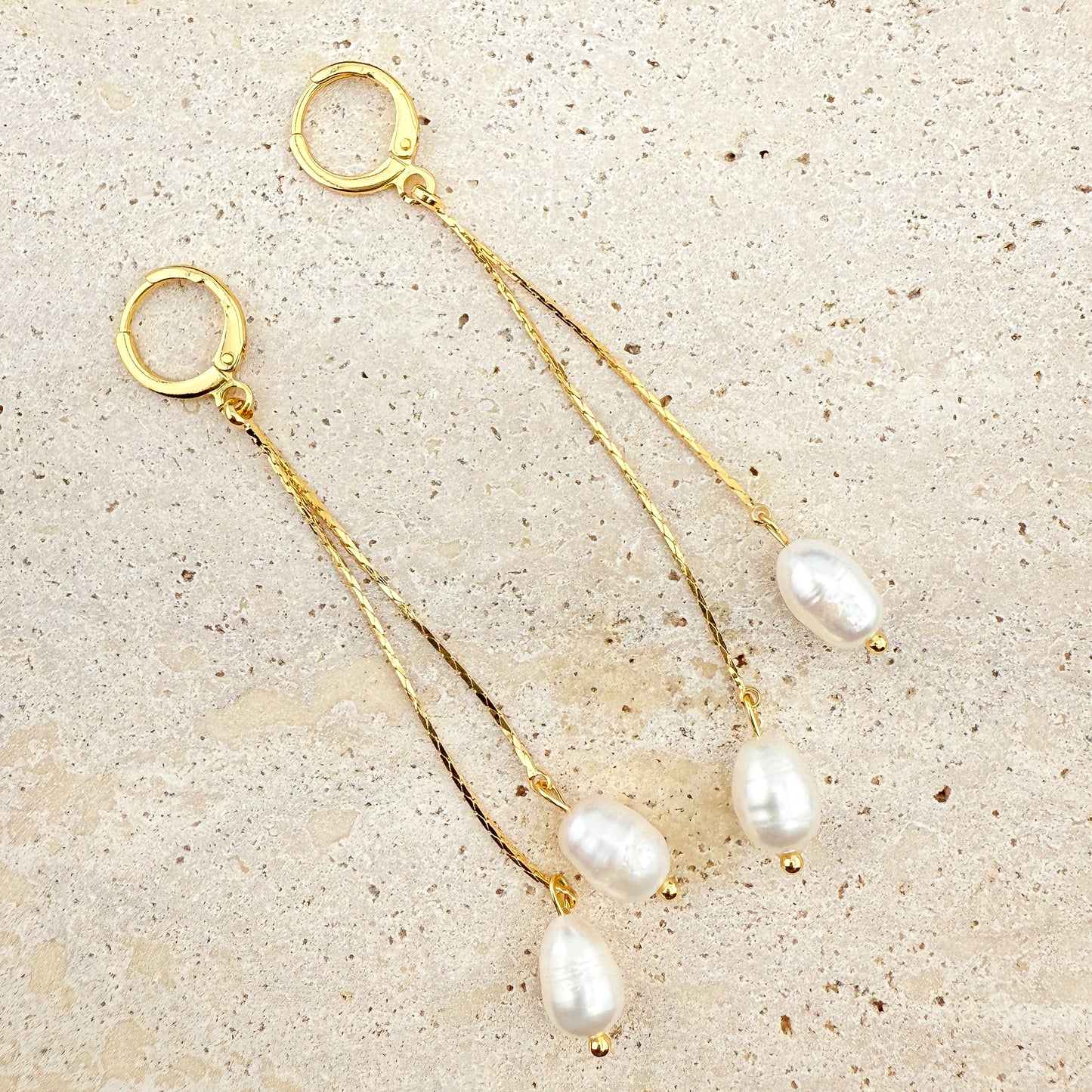 A pair of elegant Aking Sinta gold huggie hoop earrings, each featuring two delicate, dangling gold chains of varying lengths, finished with lustrous white pearls. The earrings are arranged on a light, textured natural stone background, highlighting the classic, sophisticated contrast between the polished gold and the radiant white pearls