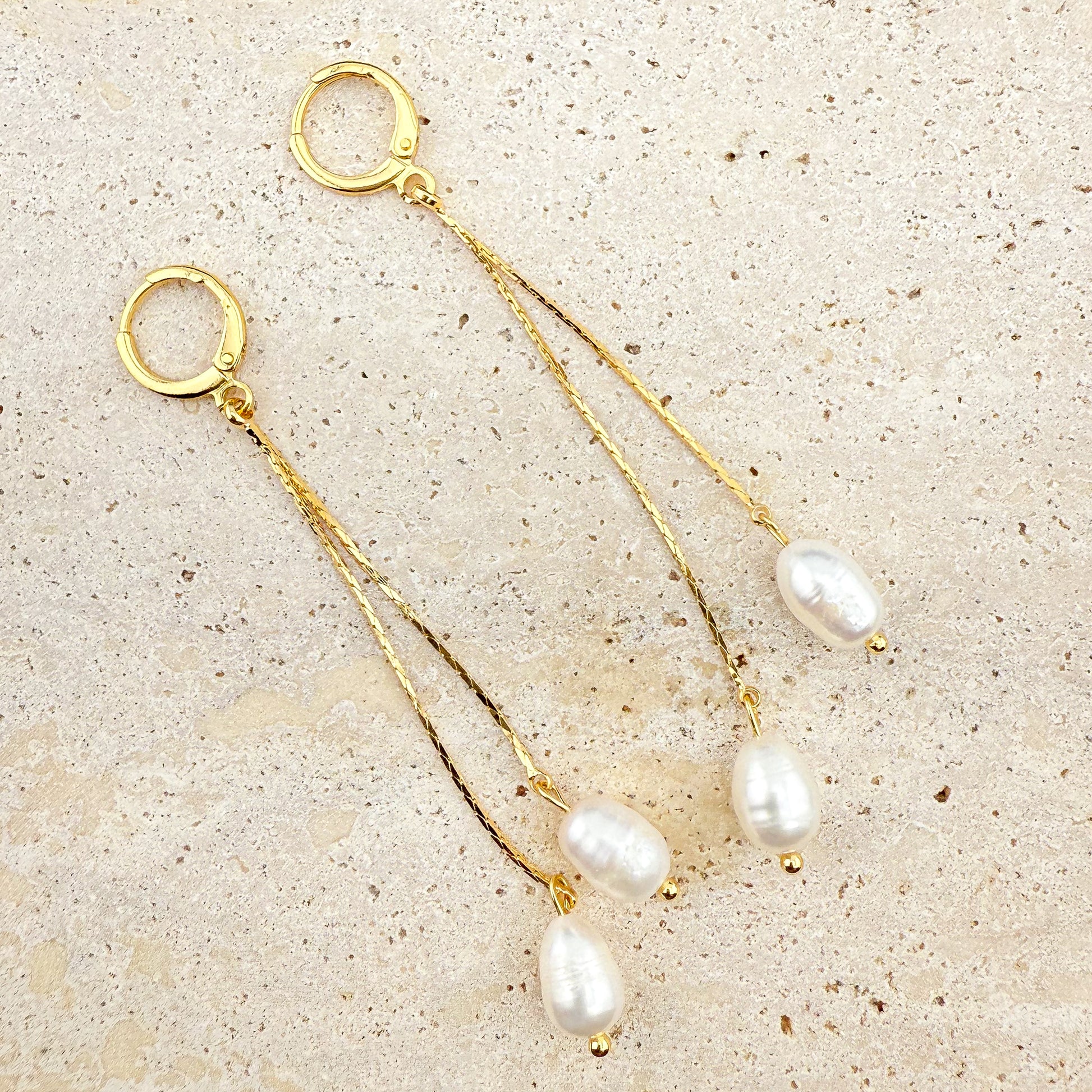 A pair of elegant Aking Sinta gold huggie hoop earrings, each featuring two delicate, dangling gold chains of varying lengths, finished with lustrous white pearls. The earrings are arranged on a light, textured natural stone background, highlighting the classic, sophisticated contrast between the polished gold and the radiant white pearls